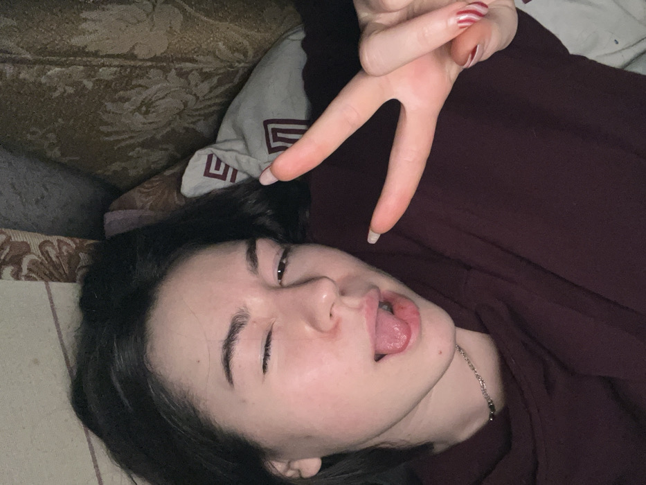 A young woman with dark hair and light skin lies down, sticking out her tongue and closing one eye. She wears a dark red shirt and a silver necklace. Her nails are long and decorated with red stripes. A hand flashes a peace sign above her face. The background includes a patterned couch, a pillow with a geometric design, and a hint of another surface.