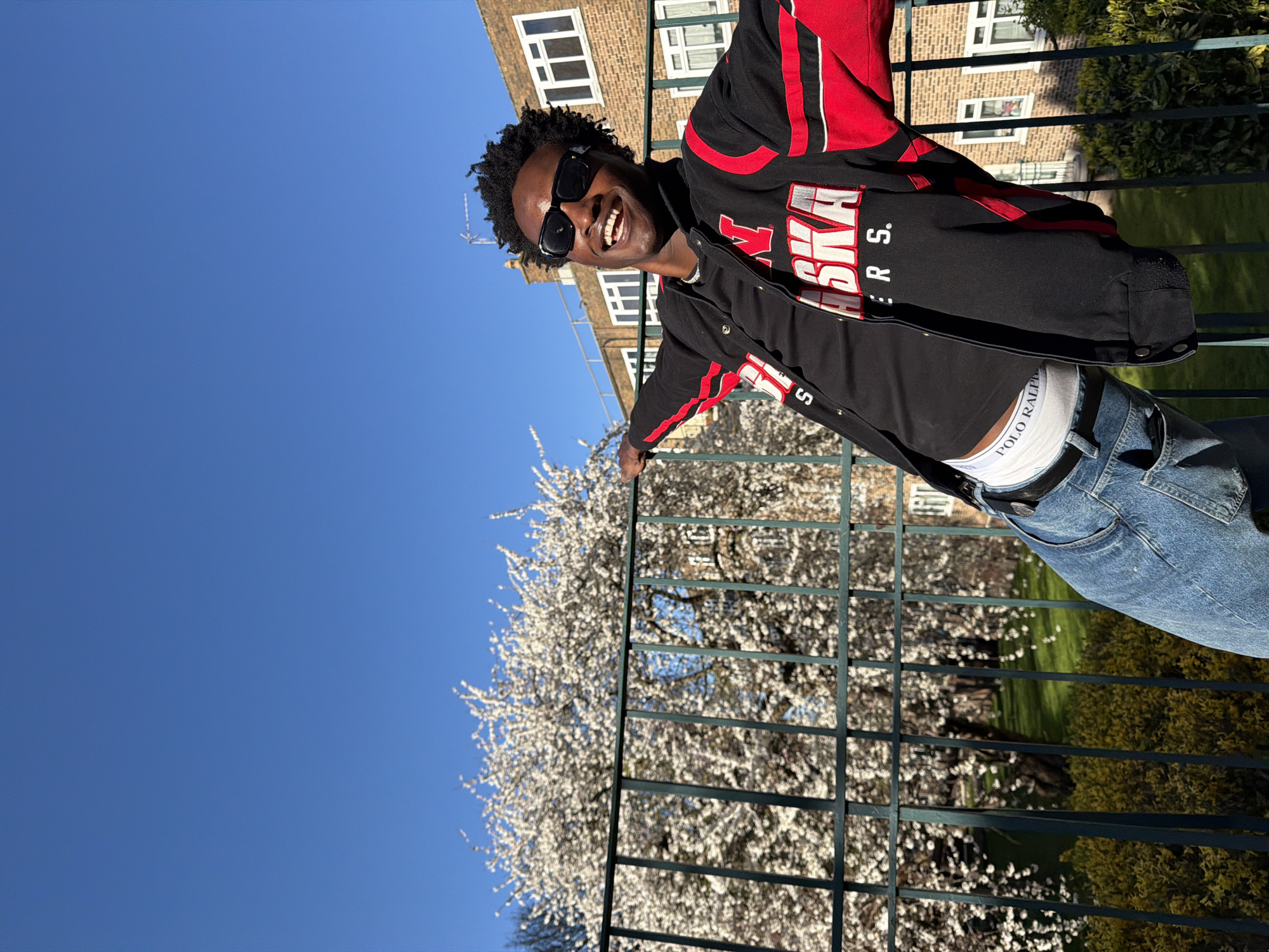 A man stands outside on a sunny day with blue skies, posing on a balcony behind a dark green metal railing with white blossoms in the background. He wears sunglasses, a black jacket with red accents and “Nebraska” printed on the front, a black t-shirt, light blue jeans, and white underwear visibly above the waistband. A building with brick and white-framed windows is visible behind him. The man appears to be laughing or smiling. While the exact location cannot be definitively determined, the architecture and environment suggest this scene could be in London, United Kingdom.