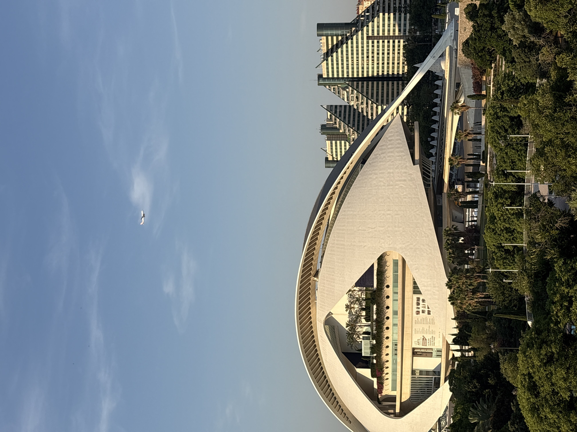 The image shows a wide-angle view of the City of Arts and Sciences in València, Spain. In the foreground, a large, white, wave-like structure, which is part of the Hemisfèric, dominates the scene. This building features a central dark void and a surrounding white canopy with diagonal louvers. Above the Hemisfèric's interior is a lush green terrace with palm trees. In the middle ground, behind the Hemisfèric, are several distinctive, triangular-shaped modern buildings with many windows and balconies, characteristic of the City of Arts and Sciences architecture. A verdant park area with mature trees and manicured shrubs occupies the lower portion of the image. In the sky, a white airplane is visible in the upper right quadrant, flying horizontally. The sky is clear and blue with some wispy white clouds.