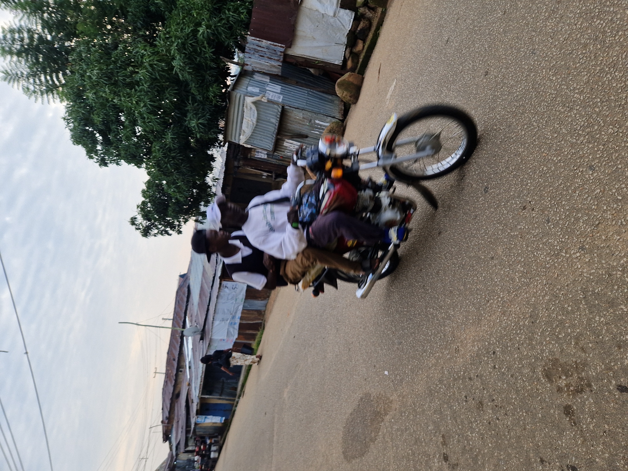 The image depicts a street scene in Jos, Nigeria, with a focus on a motorcycle carrying two individuals. The motorcycle, a red and silver model, is in motion, indicated by the blur of its wheels and the slight motion blur on the riders. The driver, wearing a white long-sleeved shirt, a white cap, and brown pants, is operating the motorcycle. Behind him, a passenger wearing a white t-shirt and a black vest with brown pants is seated. They are on a paved road with visible oil stains.
To the left of the road, several buildings with corrugated metal roofs are visible. One building has a blue facade and what appears to be a poster on its wall. Further down the street, a person is walking away from the camera, wearing a dark top and a patterned skirt, with a backpack.
To the right of the road, there is a dense green tree with lush foliage. Behind the buildings on the right, more structures made of corrugated metal and some rough-hewn wooden elements are visible. Large rocks are scattered on the ground in this area. The sky above is overcast with a light grey hue. The overall impression is of a typical street in a residential or commercial area of Jos, characterized by its architecture and the presence of everyday activities like motorcycle transport.