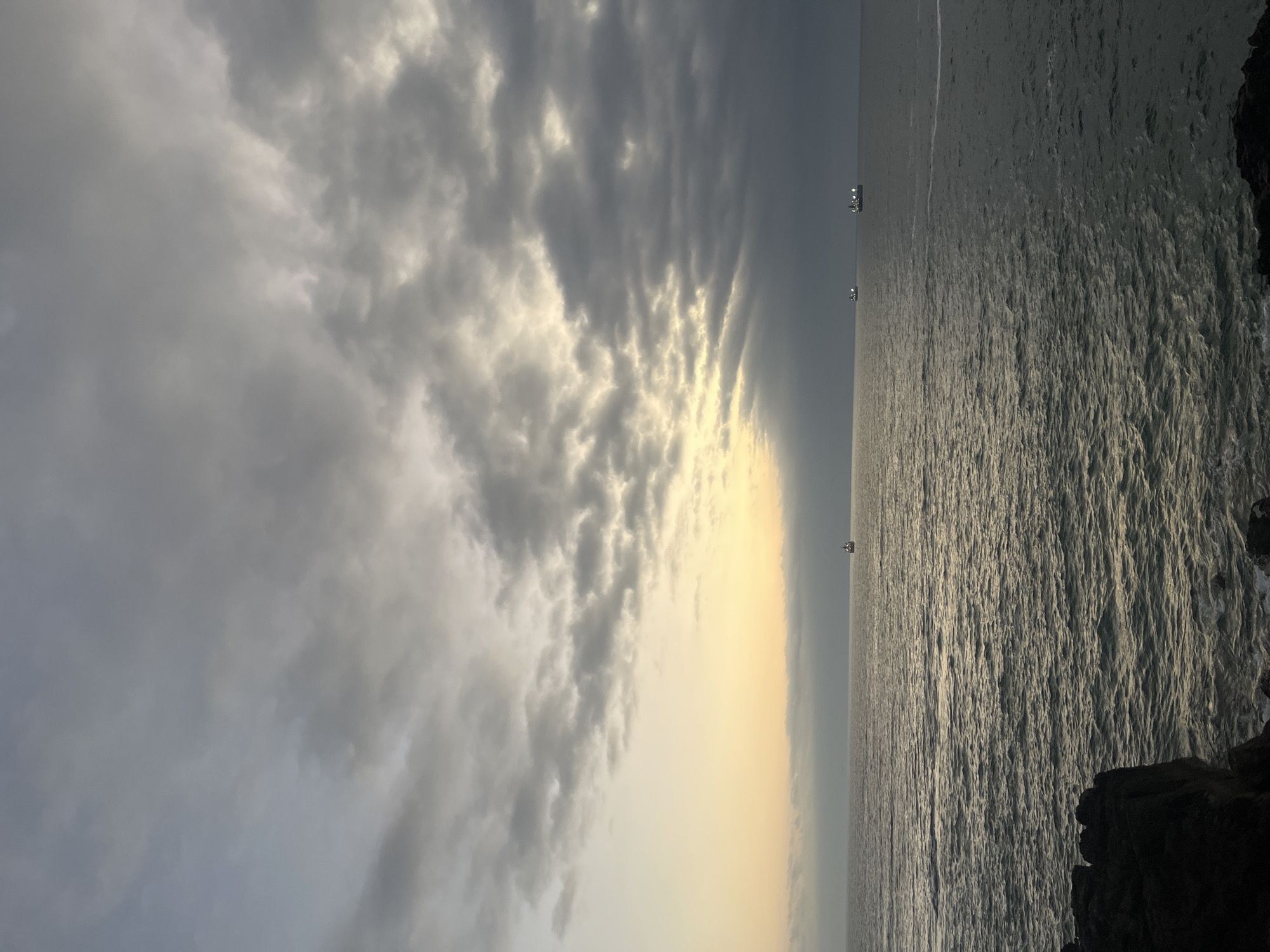 The image depicts a seascape in Arica, Chile, notable for its dynamic cloud formations and atmospheric lighting. The left side of the image is dominated by a band of grey clouds, interspersed with patches of sunlight breaking through. To the right, the ocean surface reflects the light, creating a shimmering effect. Several small boats can be seen on the horizon. Dark rocky outcroppings, likely part of the coastal geography, are visible along the right edge of the image. The horizon appears relatively flat, indicating a calm or mildly choppy sea. Overall, the scene captures a serene moment with contrasting elements of light and shadow across the sky and sea.