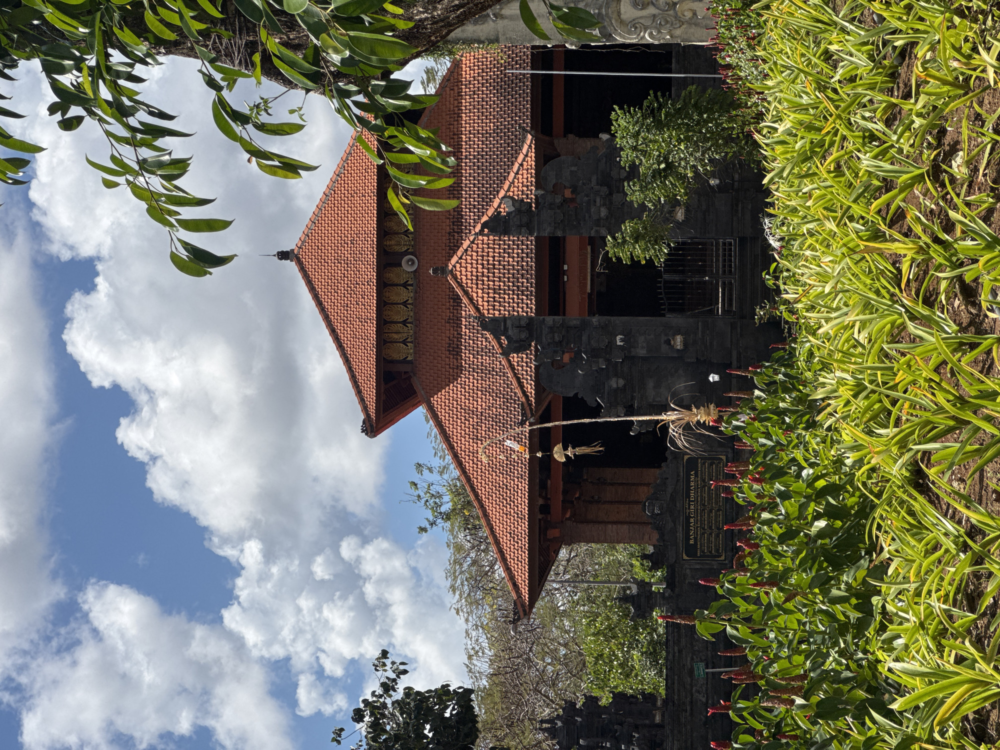 The image depicts the exterior of the Banjar Girie Dharma building, a place of worship located in Kuta Selatan, Indonesia. The building features a traditional Balinese architectural style with a prominent red-tiled roof and dark stone walls. In the foreground, a lush garden is filled with various plants, including tall, leafy green foliage and a cluster of red flowers. A signpost with Balinese script is visible in the garden. The sky above is bright blue with scattered white clouds. A large tree with green leaves is positioned on the right side of the frame, partially obscuring the building. The scene appears to be a sunny day, likely during daylight hours.