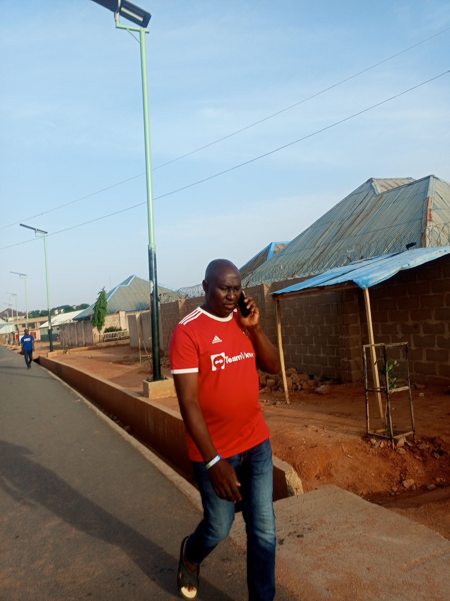 A medium shot shows a man wearing a red t-shirt with the TeamViewer logo, blue jeans, and sandals talking on a cell phone while walking on a road in Tempure, Nigeria. The road has a curb and drainage ditch to the right. A second man is walking in the distance on the same road. Streetlights line the road. Buildings are visible in the background, one with a corrugated metal roof topped with barbed wire. A makeshift shelter leans against a block wall. The ground on the right side of the road is reddish-brown dirt. The sky is light blue.
