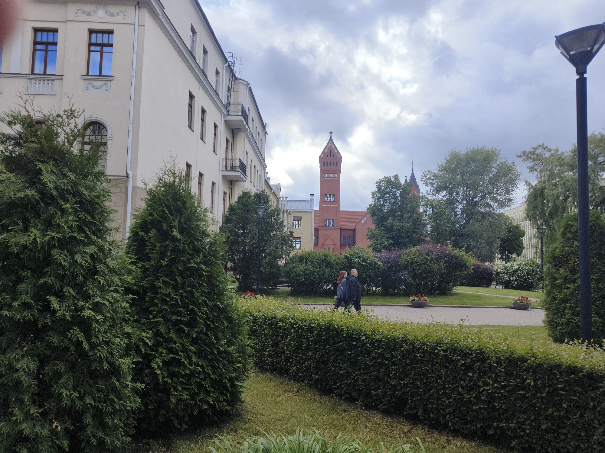 The image shows a view of Trinity Suburb in Minsk, Belarus, with the Church of the Holy Trinity and St. Elizabeth in the background. In the foreground, a manicured hedge runs diagonally across the frame, with lush green grass on either side. Two large evergreen trees are on the left, and another on the far right. In the middle ground, two people are walking on a paved pathway, with a well-kept garden area behind them. The buildings in the background are a mix of older architectural styles, with the prominent red brick church standing out. The sky is overcast, with scattered clouds.