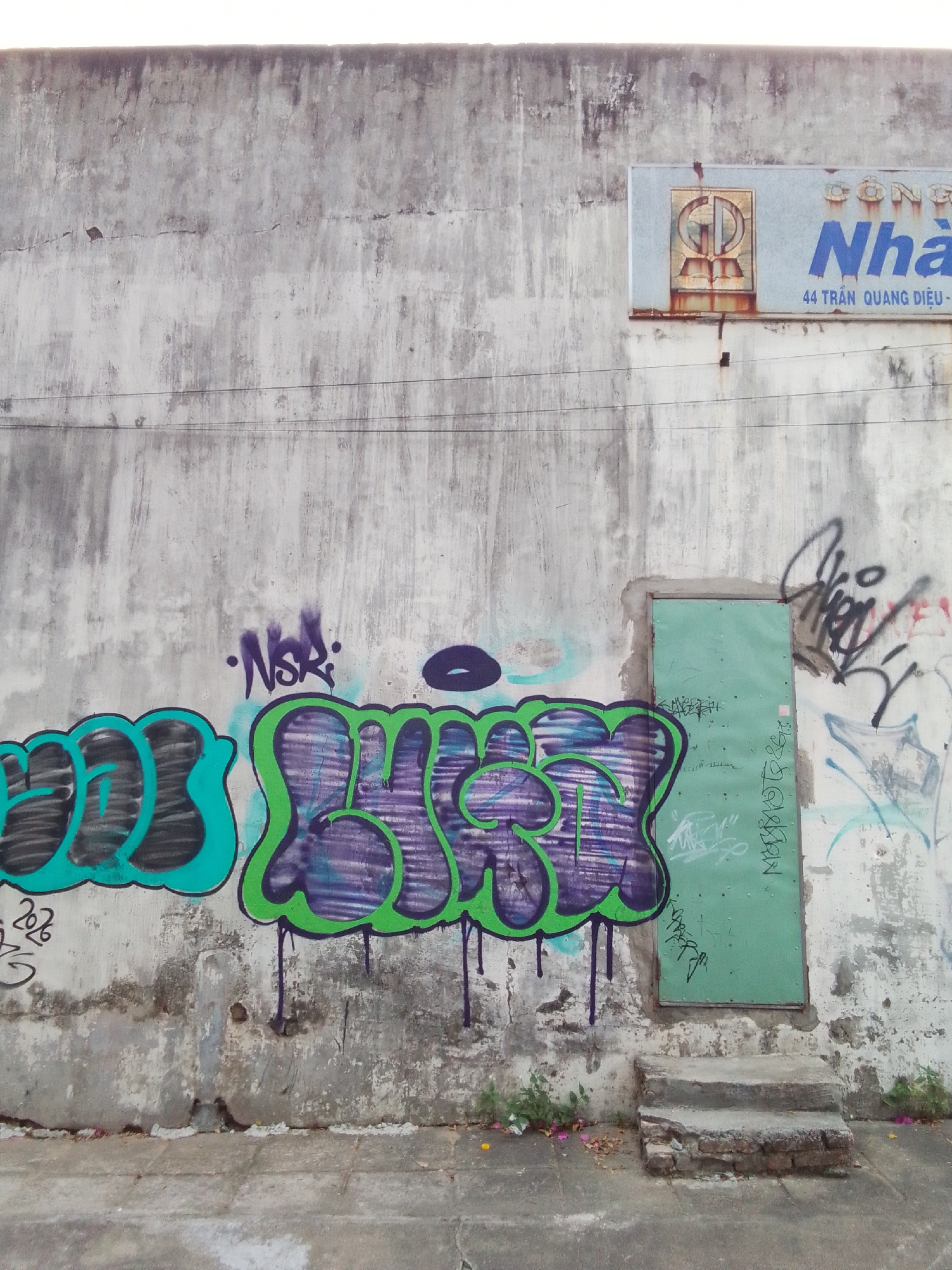 The image shows a weathered concrete wall in Da Nang, Vietnam, covered in graffiti. The wall is predominantly gray with patches of discoloration and cracks. A light blue sign with Vietnamese text and a circular logo is mounted high on the wall to the right. Below the sign, a mint green metal door is set into the wall. A small set of concrete steps leads up to the door. The lower part of the wall is adorned with large, colorful graffiti in shades of teal, purple, and green, with some black outlines and drips. Smaller tags and scribbles are visible throughout the wall, including a dark purple tag labeled "NSR". Green vines and some scattered debris are present at the base of the wall and steps. The ground in front of the wall appears to be a concrete sidewalk. The overall impression is one of urban decay and street art.