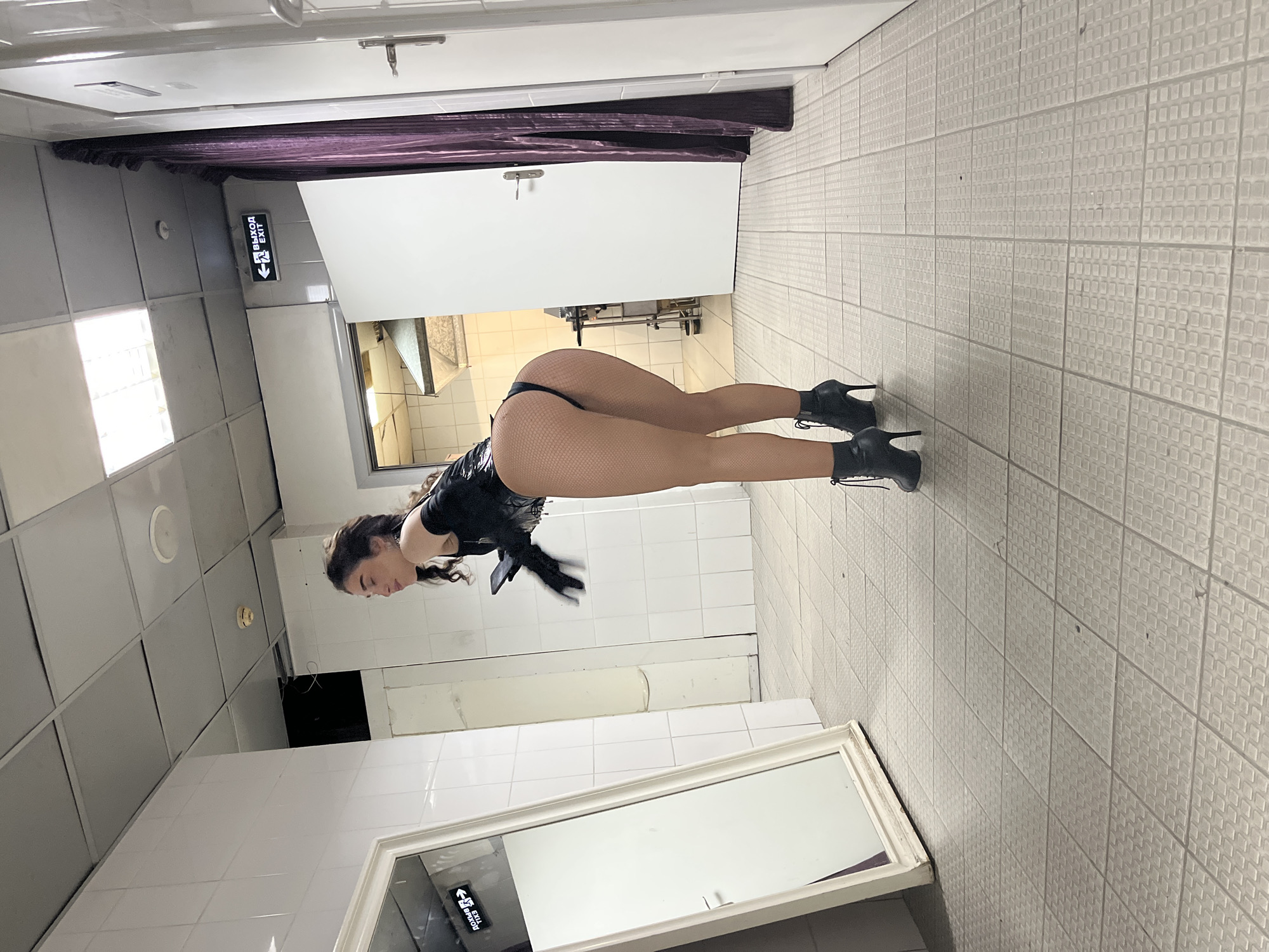 The image depicts a woman wearing a black leotard, fishnet stockings, and high-heeled boots, posing in a room with tiled walls and floors. She is bent over, with her back to the camera, and her head is turned to the side. A large mirror with a white frame is visible in the foreground, reflecting parts of the room. An exit sign with Russian text ("ВЫХОД" and "EXIT") is illuminated on the wall. The lighting suggests an indoor setting, possibly a dressing room or a backstage area. The overall mood is provocative and attention-grabbing, likely intended for artistic or performance-related purposes. The location is identified as Kazan, Russia.