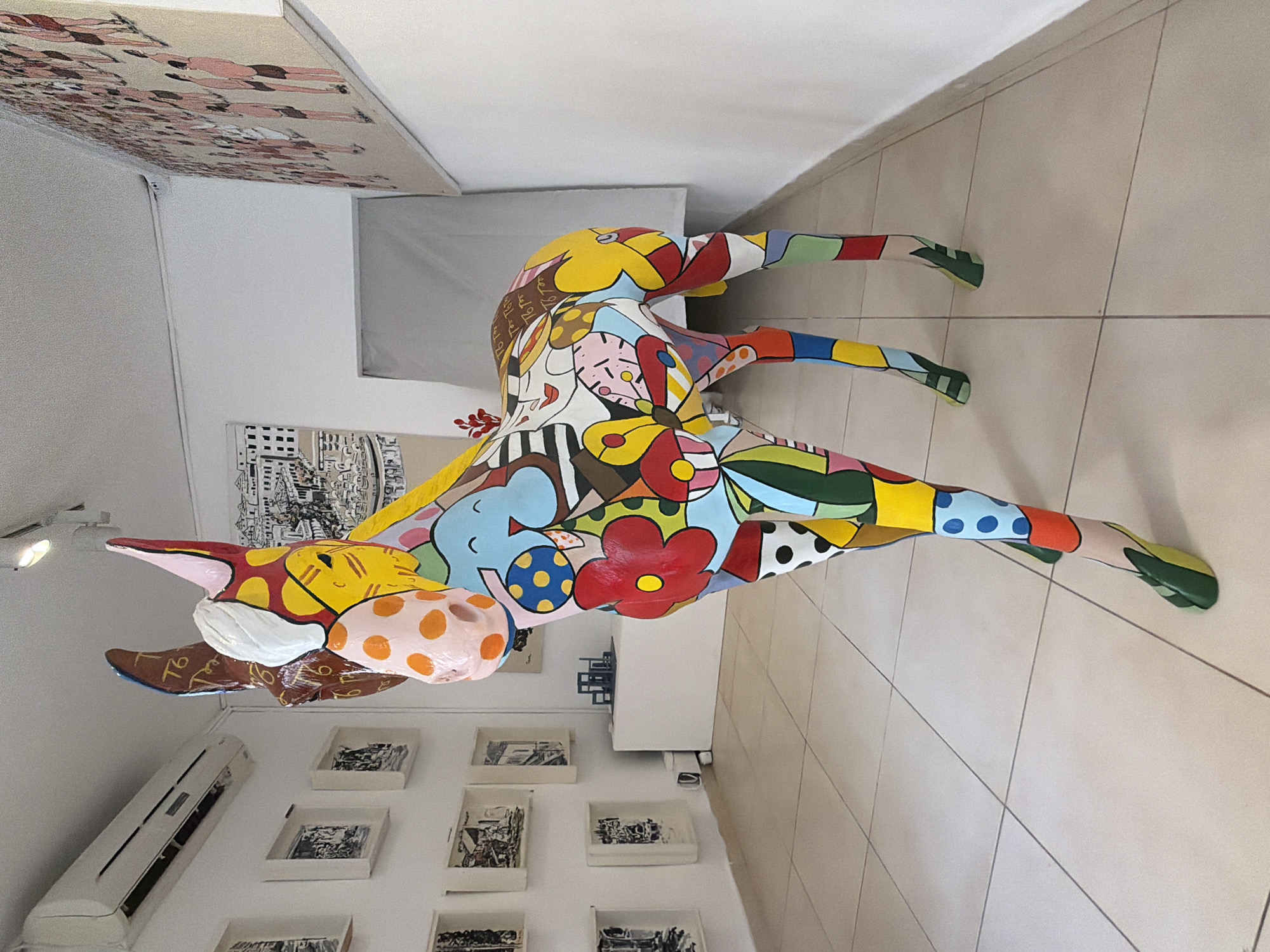 A brightly painted cow statue stands on a tiled floor. The statue has a patchwork of colors, patterns, and cartoonish elements covering its surface. It's in a room with white walls. A low wall with framed pictures is behind the statue. There is a framed piece on the ceiling. The location is likely Tel-Aviv, Israel.