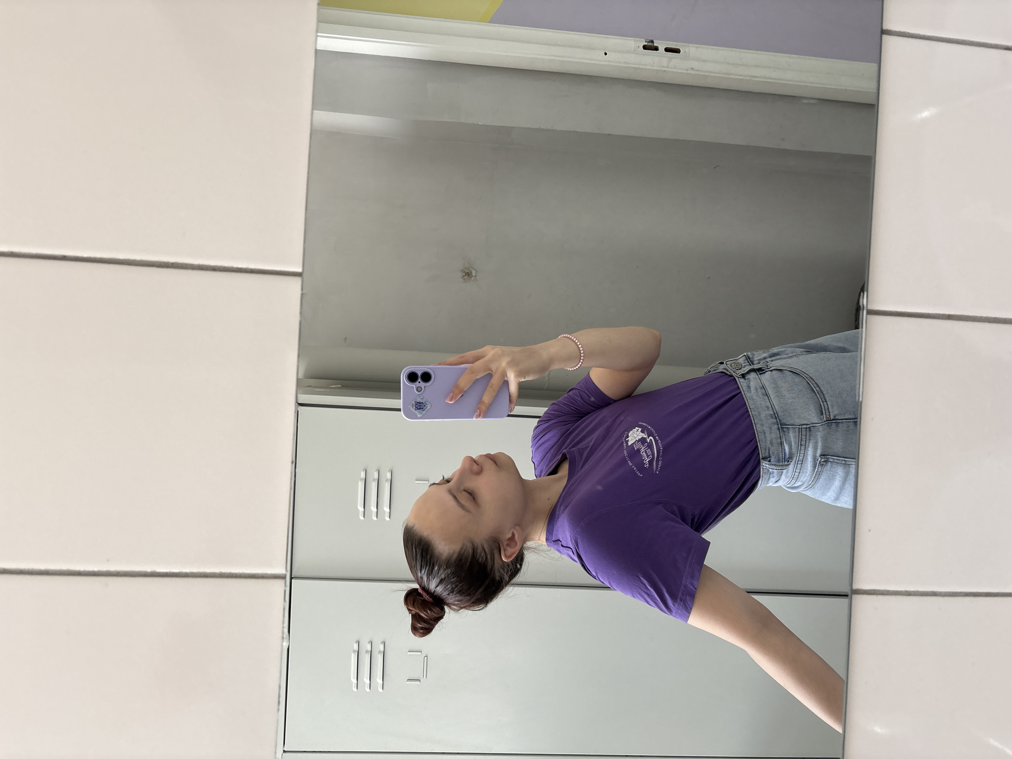 A woman is taking a mirror selfie in a room lined with white tiles on one side and what appears to be gray metal lockers on the back wall. The upper portion of the back wall features a horizontal white beam with a lavender stripe above it. The woman is wearing a purple t-shirt and denim shorts. She is holding a purple phone in her right hand. The logo on her shirt is of a company or organization in Chaikovsky, Russia.