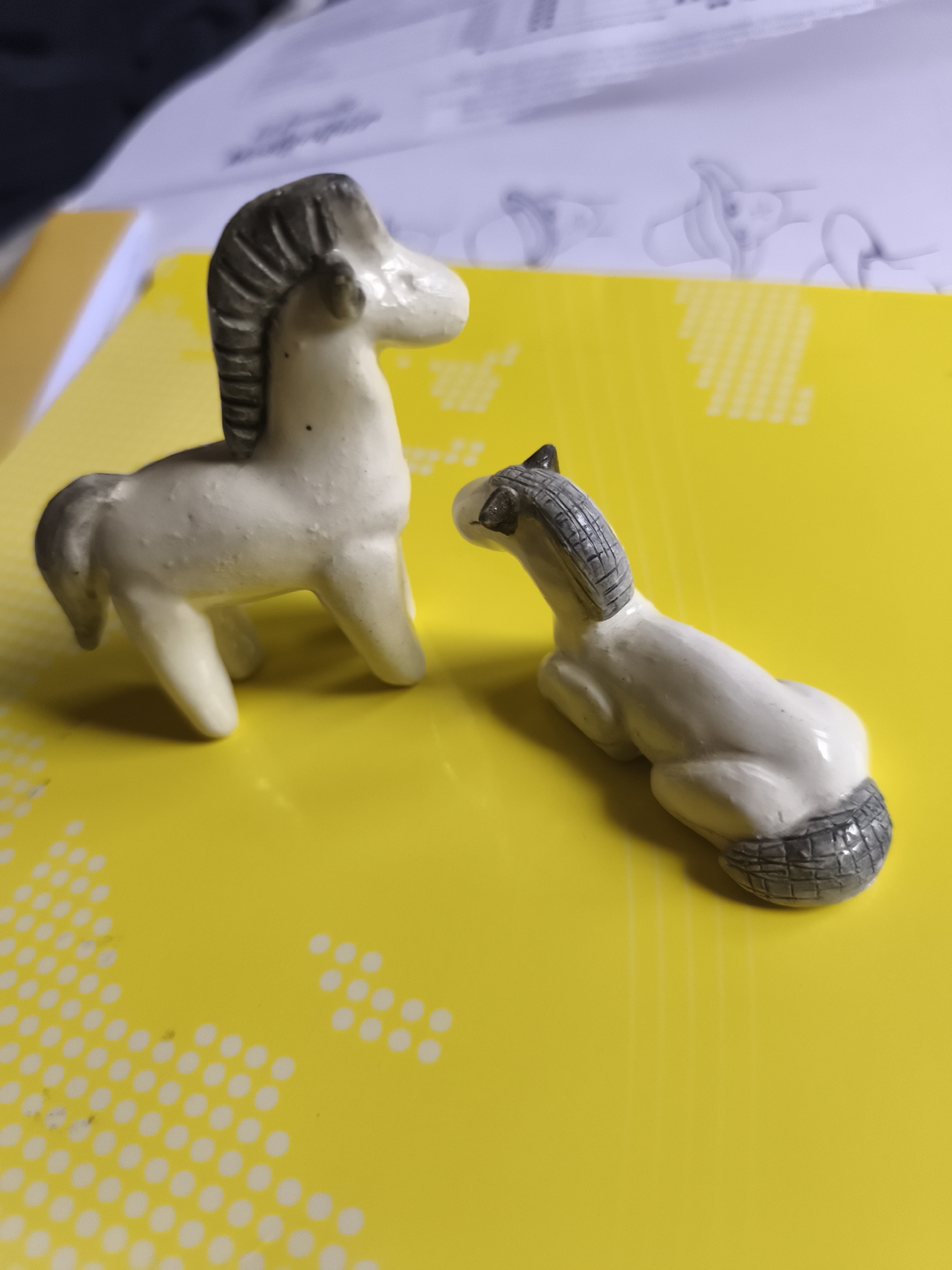 Two small, white ceramic horse figurines are positioned on a bright yellow surface with a white dotted pattern. The horse on the left is standing, facing left, with its mane and tail detailed in gray. The horse on the right is lying down, facing right, with its mane and tail also in gray. The background consists of a blurred document with text and illustrations, and a portion of a dark surface. The scene appears to be indoors, on a desk or table. There is no indication of the location in Saint-Denis, France.