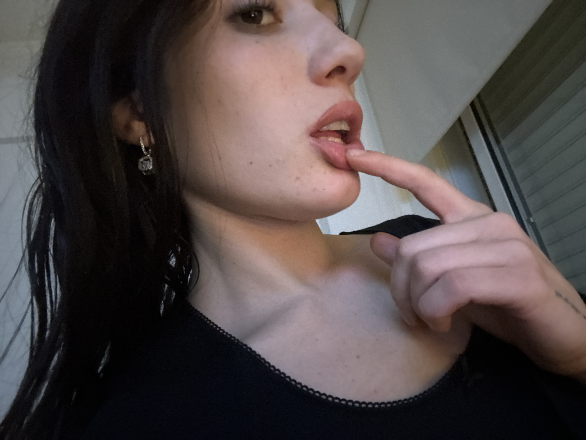 A woman is positioned in the foreground, looking off to the right of the frame with her finger touching her lower lip. She has fair skin, dark hair, and is wearing a black top with a decorative neckline. She has a small earring on her left ear. The background features a wall and a partially visible window with blinds. The location is not discernible as Buenos Aires, Argentina, based on the image provided.