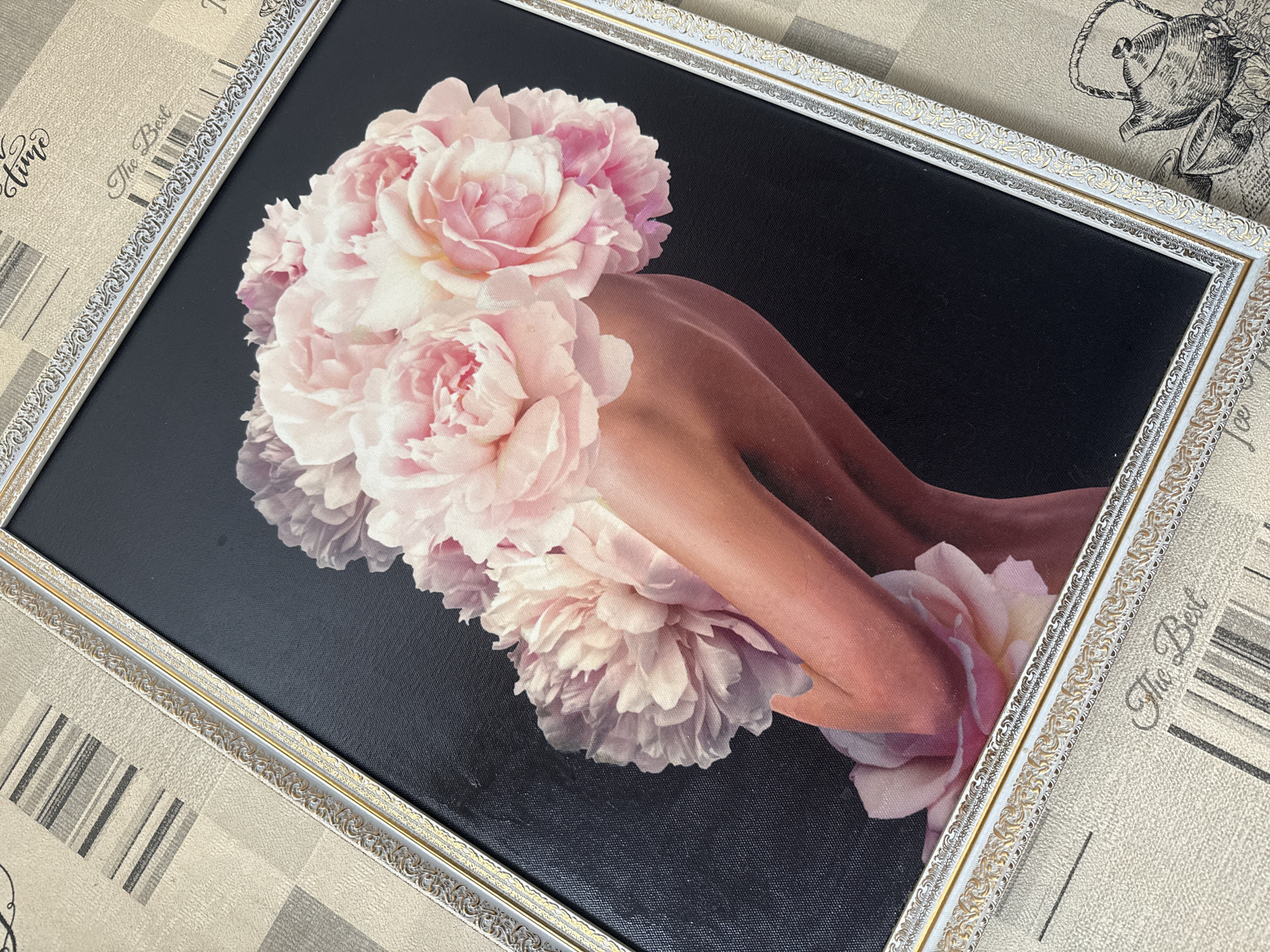 A framed artwork featuring a stylized depiction of a woman's torso emerging from a bouquet of pink peonies against a black background is displayed on a patterned surface. The frame is ornate, with white and gold accents. The background surface exhibits a repeating pattern that includes faint text, such as "The Best" and "Time," and vertical stripes. The image is shot from a slightly elevated angle, with the artwork tilted. There is no discernible information linking this image to the city of Simferopol, Ukraine.
