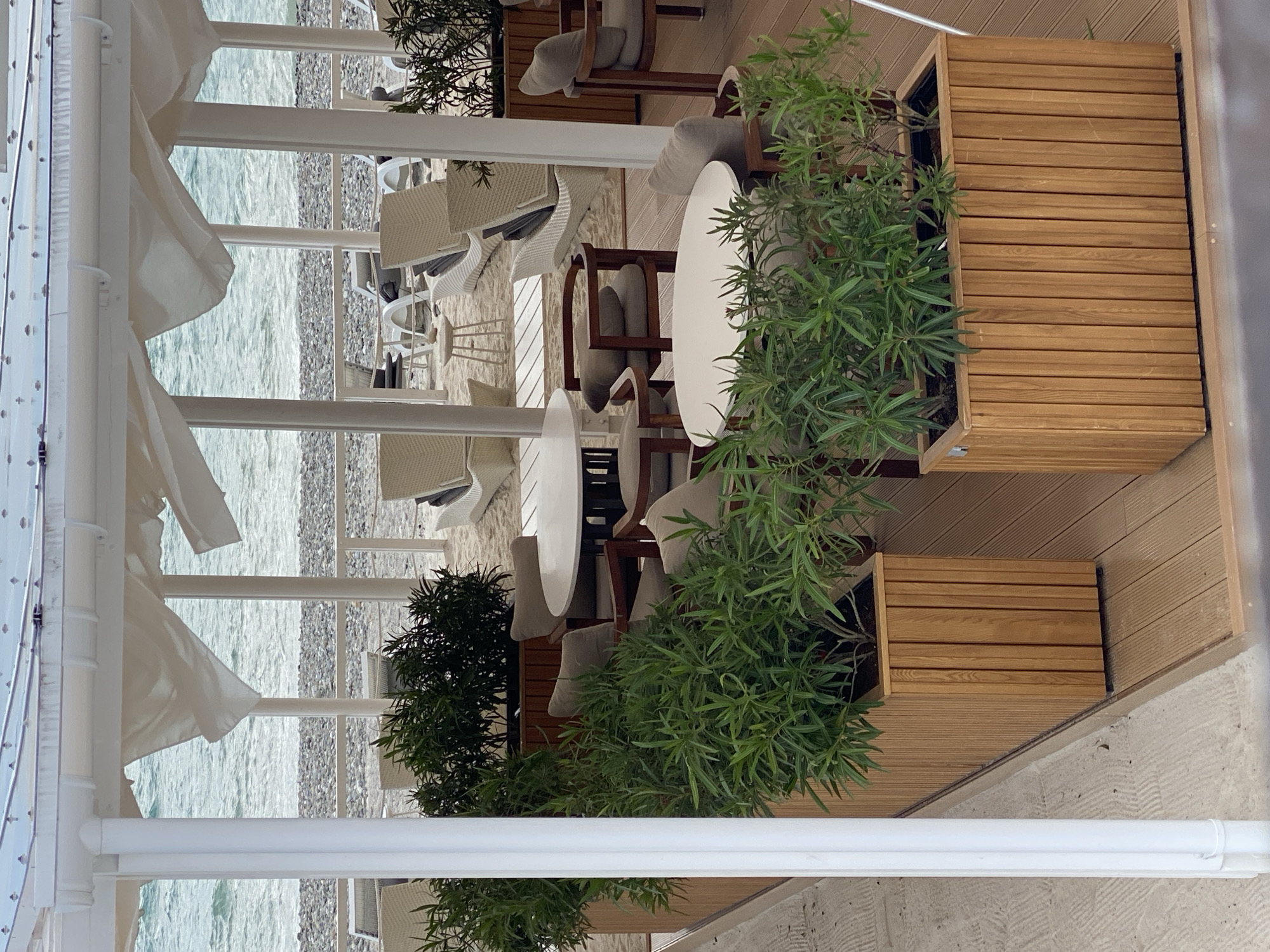 The image shows an outdoor seating area with tables and chairs. The tables are round and white, surrounded by wooden chairs with beige cushions. The seating area is located on a wooden deck with planters. The view from the seating area is of the beach and the sea. The scene is covered by a white awning. The image is shot from a high angle, looking down on the seating area. The location of the scene is in Sirius, Russia.