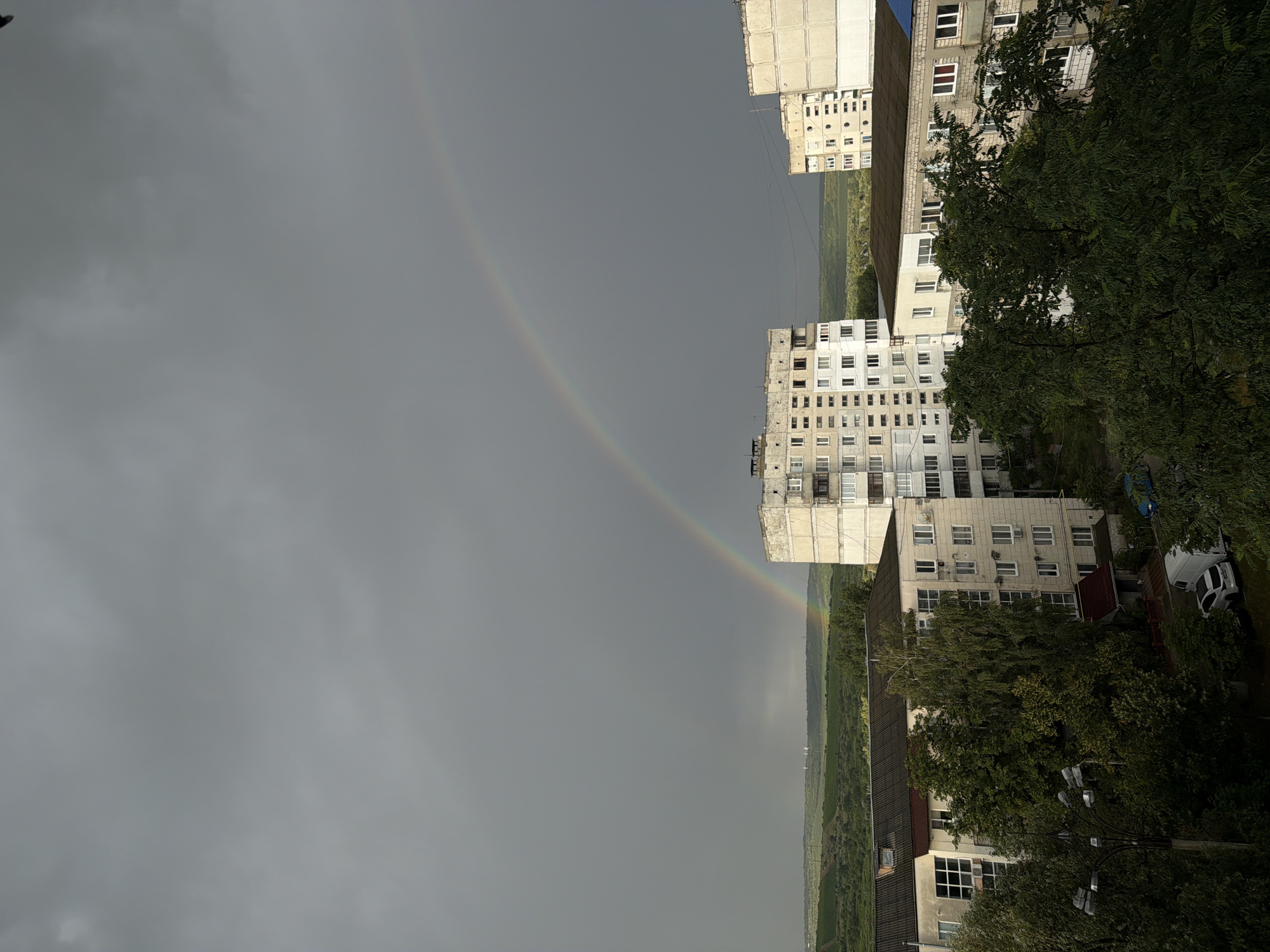 The image depicts a residential area in the city of Strășeni, Moldova, under a cloudy sky with a rainbow arcing across it. In the foreground, several multi-story apartment buildings are visible. These buildings are characterized by their concrete construction, rows of windows, and balconies. Lush green trees and foliage are interspersed among the buildings, obscuring some of the lower levels. A lamppost is also present in the left foreground. In the background, a rolling landscape of green hills and fields stretches towards the horizon, with a few distant structures or wind turbines. The overall impression is of a post-Soviet urban environment, likely captured after or during a rain shower. There are no people visible in the image. Several vehicles, including a white van and a dark-colored car, are parked near the buildings.