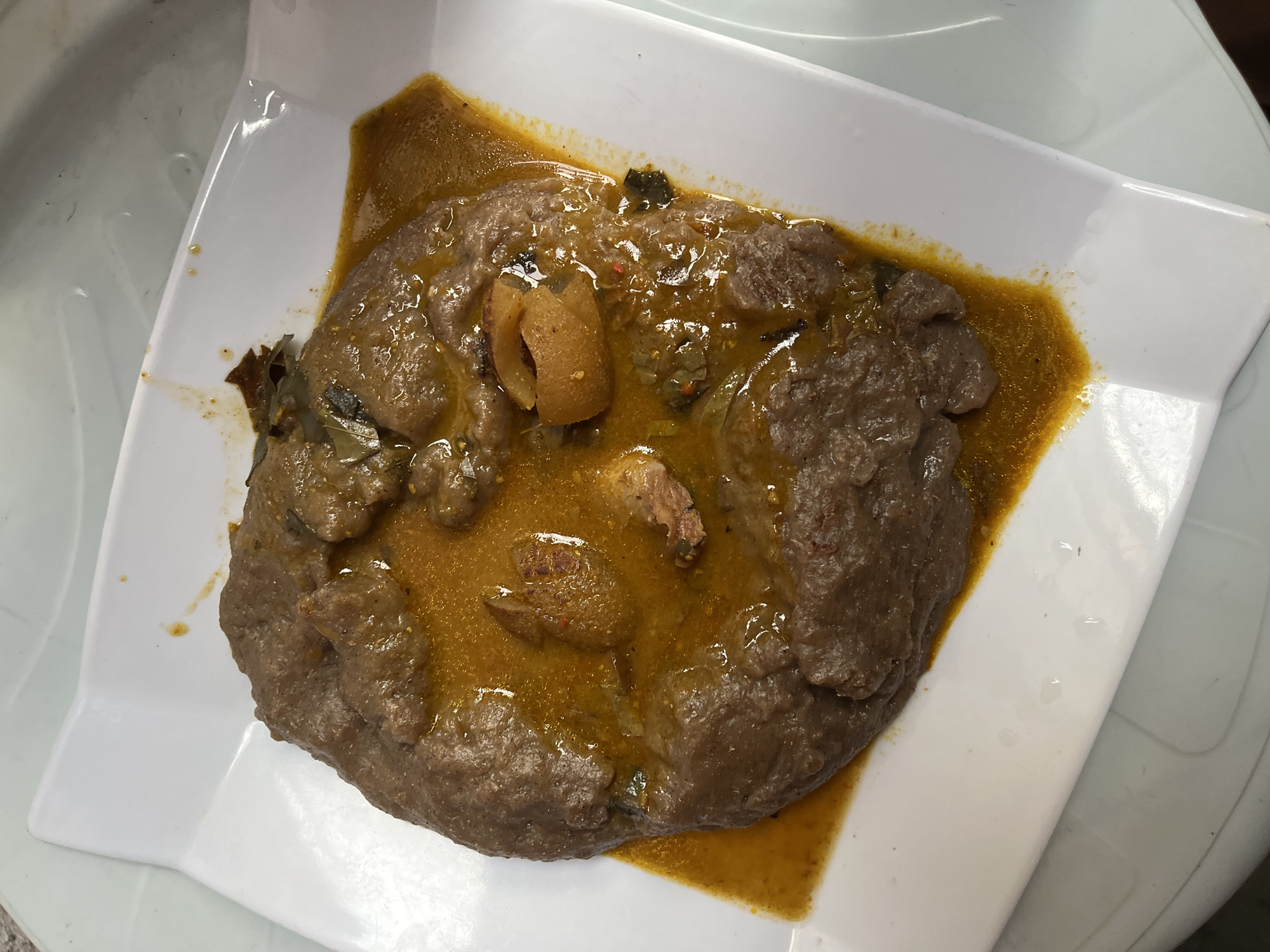 The image presents an overhead shot of a traditional Nigerian meal served on a white, rectangular plate. The central subject is a generous portion of dark brown, thick "swallow" food, likely Amala, which has a slightly rough texture. This is accompanied by a vibrant, orange-brown stew or soup, which covers a significant part of the "swallow" and pools around it.
Within the stew, various pieces of protein are visible. These include several chunks of brownish meat and a distinct yellowish, curled piece that appears to be 'ponmo' (cow skin), a common ingredient in Nigerian cuisine. Traces of green leafy vegetables and possibly red pepper flakes are also discernible in the rich, oily stew.
The white plate has gently raised edges and a somewhat reflective surface, suggesting it might be made of melamine or plastic. The background shows a light-colored, possibly plastic table or surface, with faint patterns and reflections, indicating an indoor setting.
The image captures the meal as if it's freshly served and ready for consumption. No individuals are present in the frame, and no specific event beyond the meal itself is depicted. There is no visible text. The dish's components strongly align with typical Nigerian culinary practices, consistent with the given location context of Iranla, Nigeria.