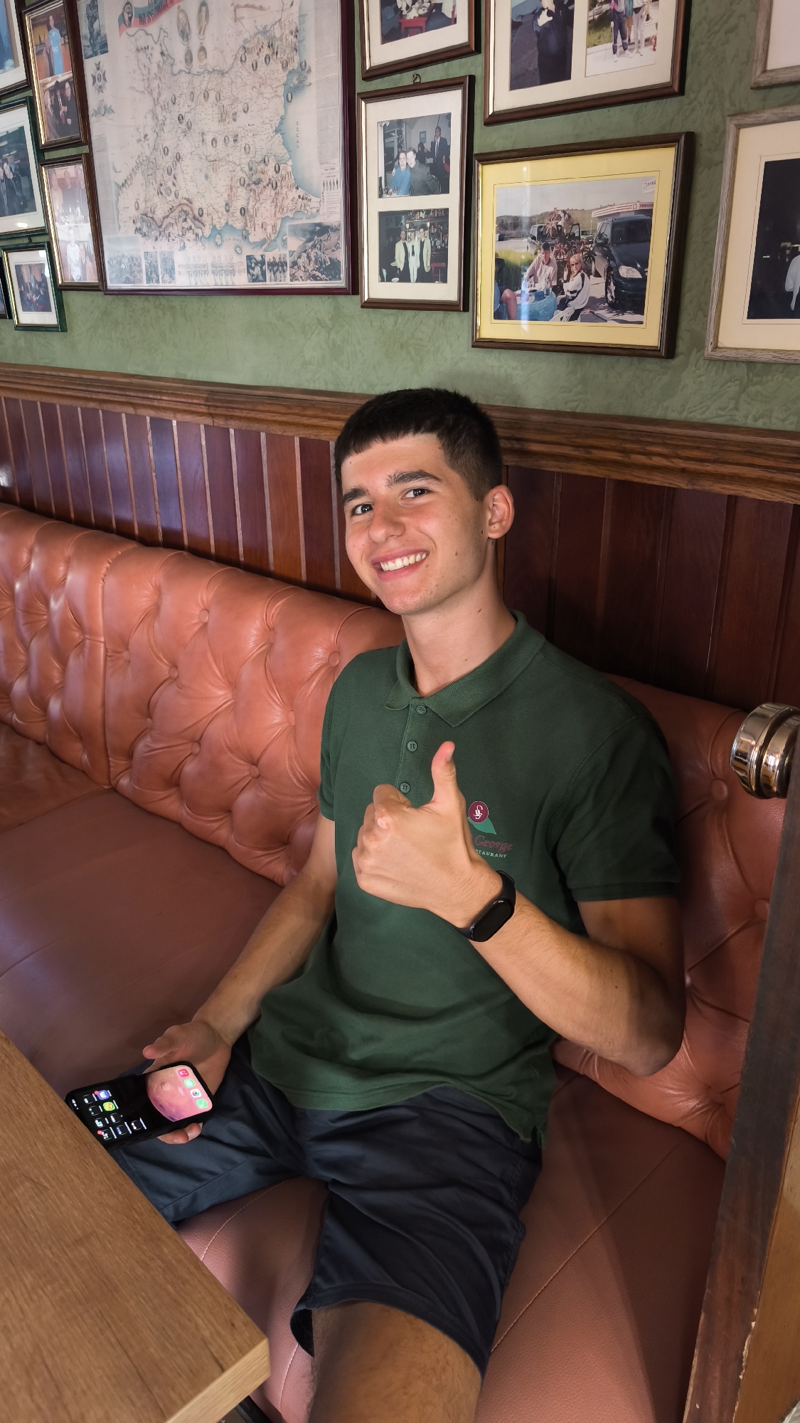 A young man with dark hair and a bright smile is seated on a tufted leather booth, giving a thumbs-up gesture. He is wearing a dark green polo shirt with a logo that reads "George's Restaurant" and dark shorts. He holds a smartphone in his left hand. The background features a wall adorned with framed photographs and a map. The ambiance suggests a restaurant setting, possibly in Pomorie, Bulgaria, given the presence of a map that appears to depict the region. The lighting is warm and inviting, contributing to a relaxed atmosphere.