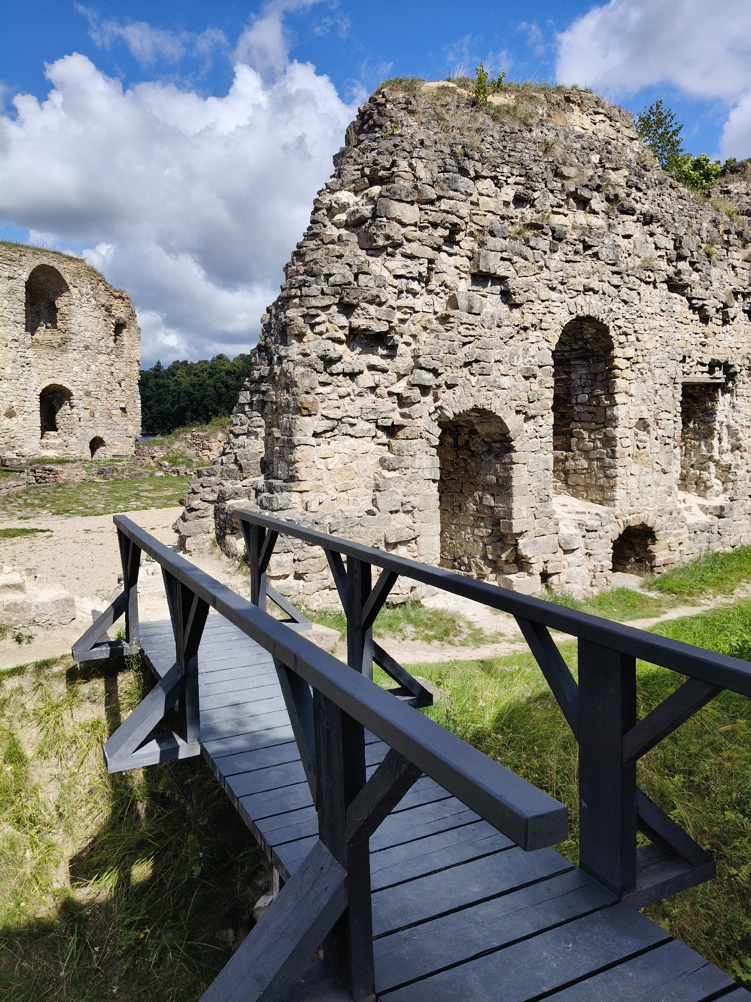 This image depicts the ruins of Koknese Castle in Koknese, Latvia. The ruins, constructed from large, rough-hewn stones, dominate the frame, with a large, prominent section of the castle wall in the foreground and to the right, featuring several arched openings. Another section of the castle, appearing as a tower-like structure, is visible in the background to the left, also with arched windows. A simple wooden footbridge, painted dark grey, spans the foreground, leading towards the ruins. The bridge has sturdy railings made of dark wood. The ground around the ruins consists of dry, sandy soil and patches of green grass and vegetation. The sky above is a bright blue with scattered, fluffy white clouds. The overall scene conveys a sense of historical decay and preservation under a clear, sunny sky. There are no people visible in the image.