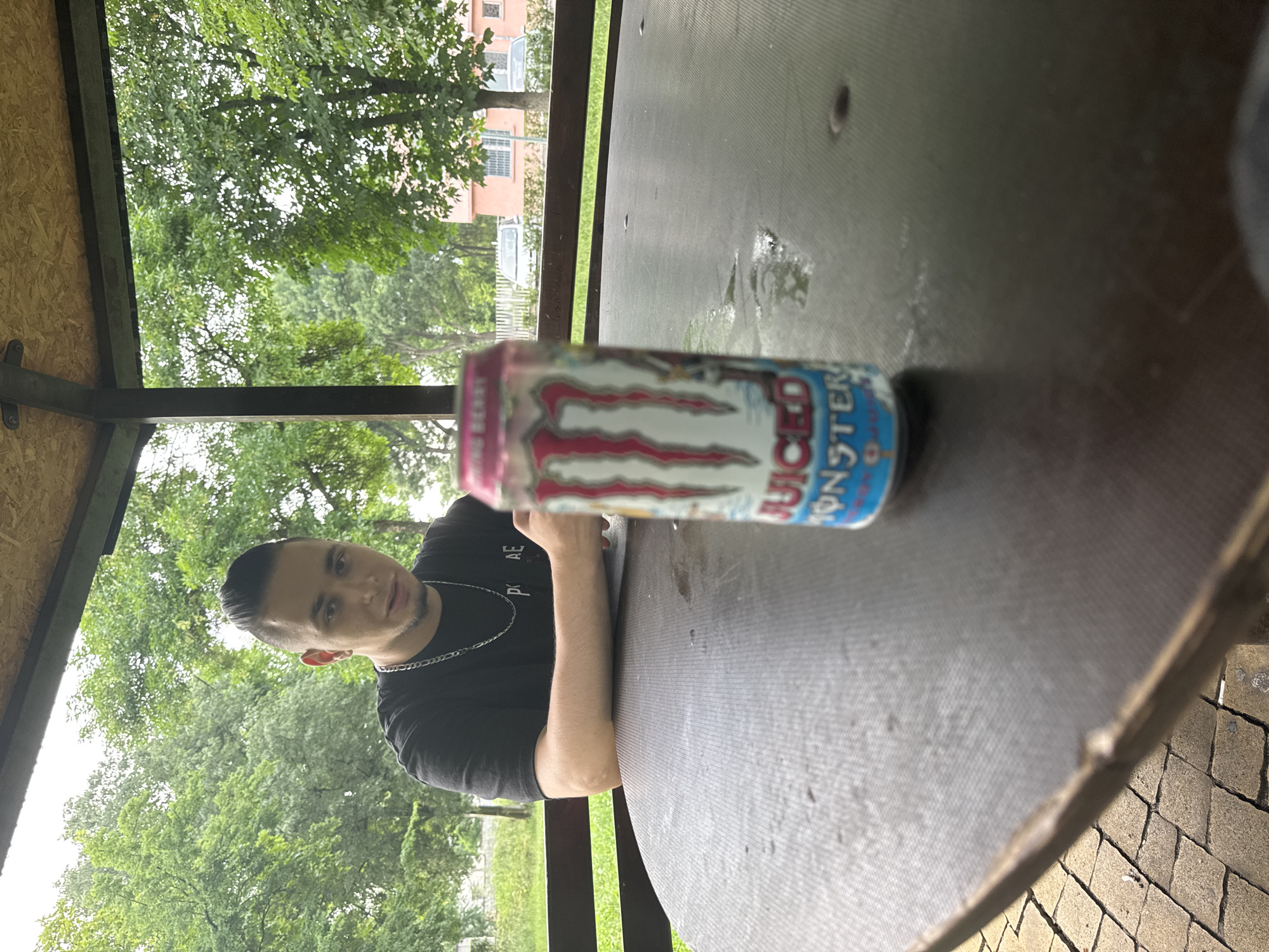 The image, appearing rotated 90 degrees counter-clockwise, captures a young man and an energy drink can within an outdoor shelter, likely a gazebo or pergola, in Radnevo, Bulgaria.
The subject is a man, positioned in the lower-left, with short dark hair, stubble, and wearing a black t-shirt with a silver chain necklace. He is leaning on a dark, textured table with his left arm visible and looks directly towards the camera with a neutral expression.
A prominent object is a Monster "Juiced" energy drink can, lying on its side on the table in the foreground. The can is colorful with pink, white, and red tones, featuring the recognizable Monster claw logo. The words "JUICED" and "MONSTER" are clearly visible, as is a partial flavor name, possibly "KING BERRY," on the left side of the can.
The scene is set outdoors, characterized by a lush green background of trees and foliage, suggesting a park or recreational area. Through the greenery, parts of light-colored buildings can be seen. The shelter features a dark grey/brown table surface, dark support beams, and light wooden ceiling panels (possibly OSB). The ground visible in the bottom right corner appears to be paved with cobblestones. The lighting is soft and diffused, indicative of an overcast day or a well-shaded area. The activity appears to be the man relaxing or taking a break.
