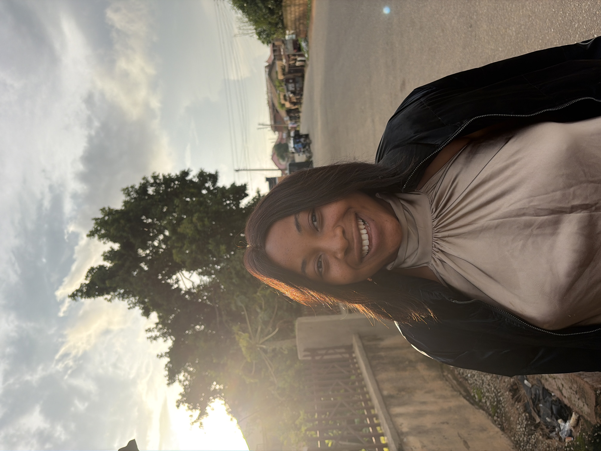 A medium shot captures a Black woman with dark hair smiling at the camera, standing outdoors in what appears to be a residential area in Jos, Nigeria. She is wearing a light beige, sleeveless top with a high neckline and a black, zip-up jacket. The sun is setting behind her, casting a warm glow and creating lens flare from the left side of the frame. The background shows a street lined with buildings and trees. The road appears to be paved and there are some power lines visible. The overall scene suggests an informal, everyday moment captured during the golden hour.