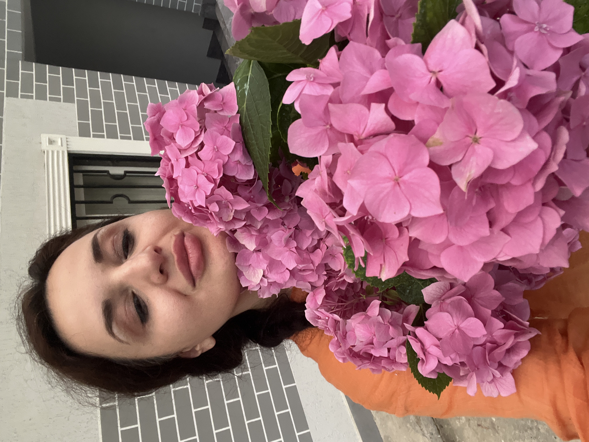 A woman is holding a large bouquet of pink hydrangeas in front of her face. The woman has dark hair and is wearing an orange top. Her eyes are closed, and her lips are slightly parted, giving her a relaxed and contented expression. The hydrangeas are vibrant pink and fill the right side of the frame, obscuring much of the woman's face. The background appears to be an outdoor setting with a wall of gray bricks and white grout. A window with a black grate is visible behind the flowers. The lighting suggests it could be daytime.