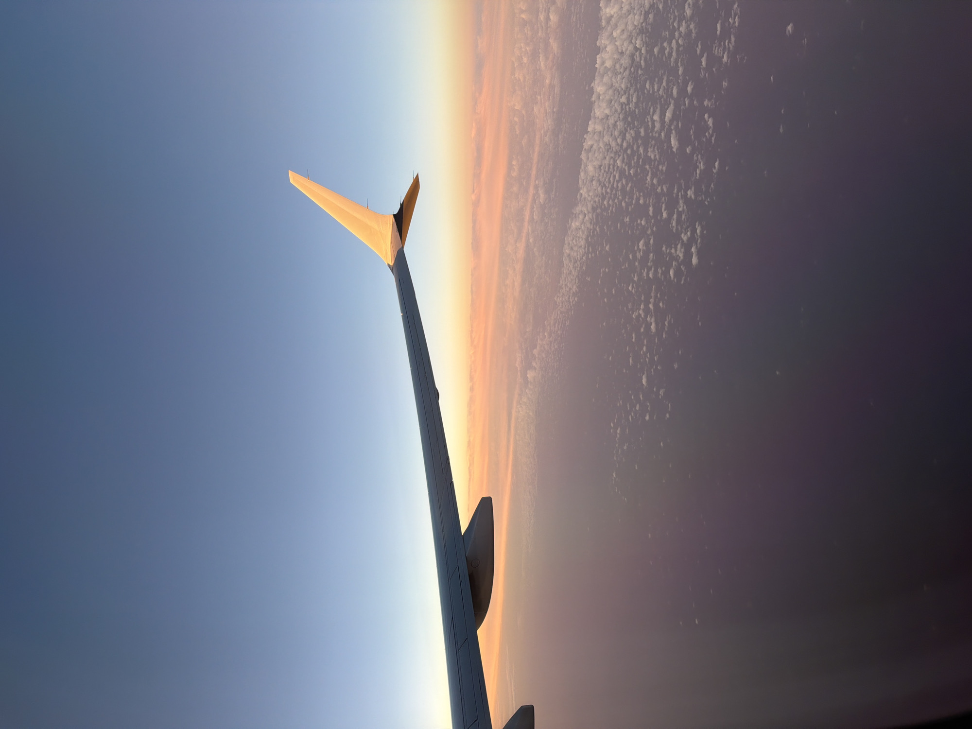 A tilted, wide shot from an airplane window features the aircraft's wing against a backdrop of blue sky and a cloudy horizon. The wing is prominently visible, spanning from the bottom to the top center, with its wingtip angled towards the upper left corner. The lighting suggests either sunrise or sunset, casting a warm glow on the clouds along the horizon. The sky transitions from a lighter blue near the horizon to a darker shade further away. The clouds are dense and textured, fading into darkness at the bottom of the frame. The scene is devoid of any ground features, focusing solely on the aerial view.