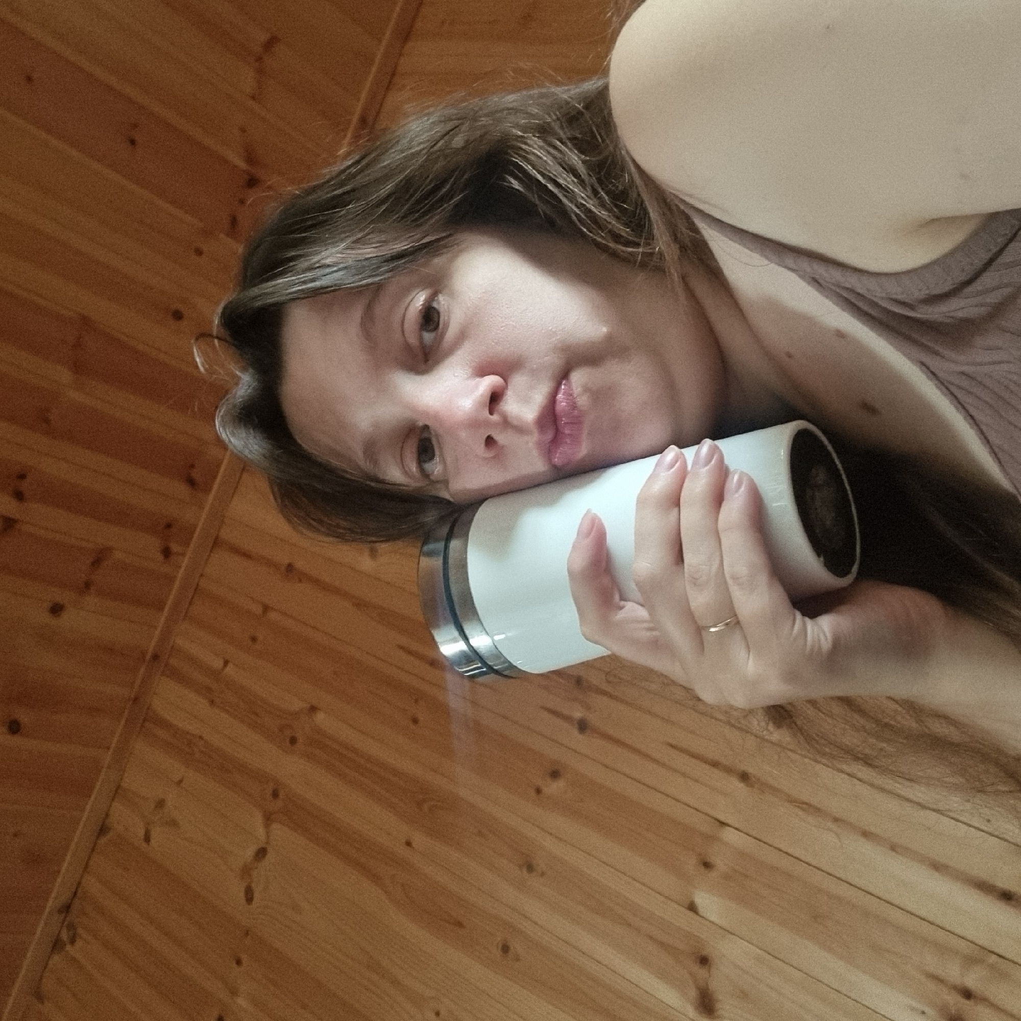 The image shows a close-up of a woman holding a white insulated mug to her cheek. She has brown hair and is wearing a brown tank top. The background is a wooden paneled wall. There is no visible indication of the location being in Dunnae-myeon, South Korea.