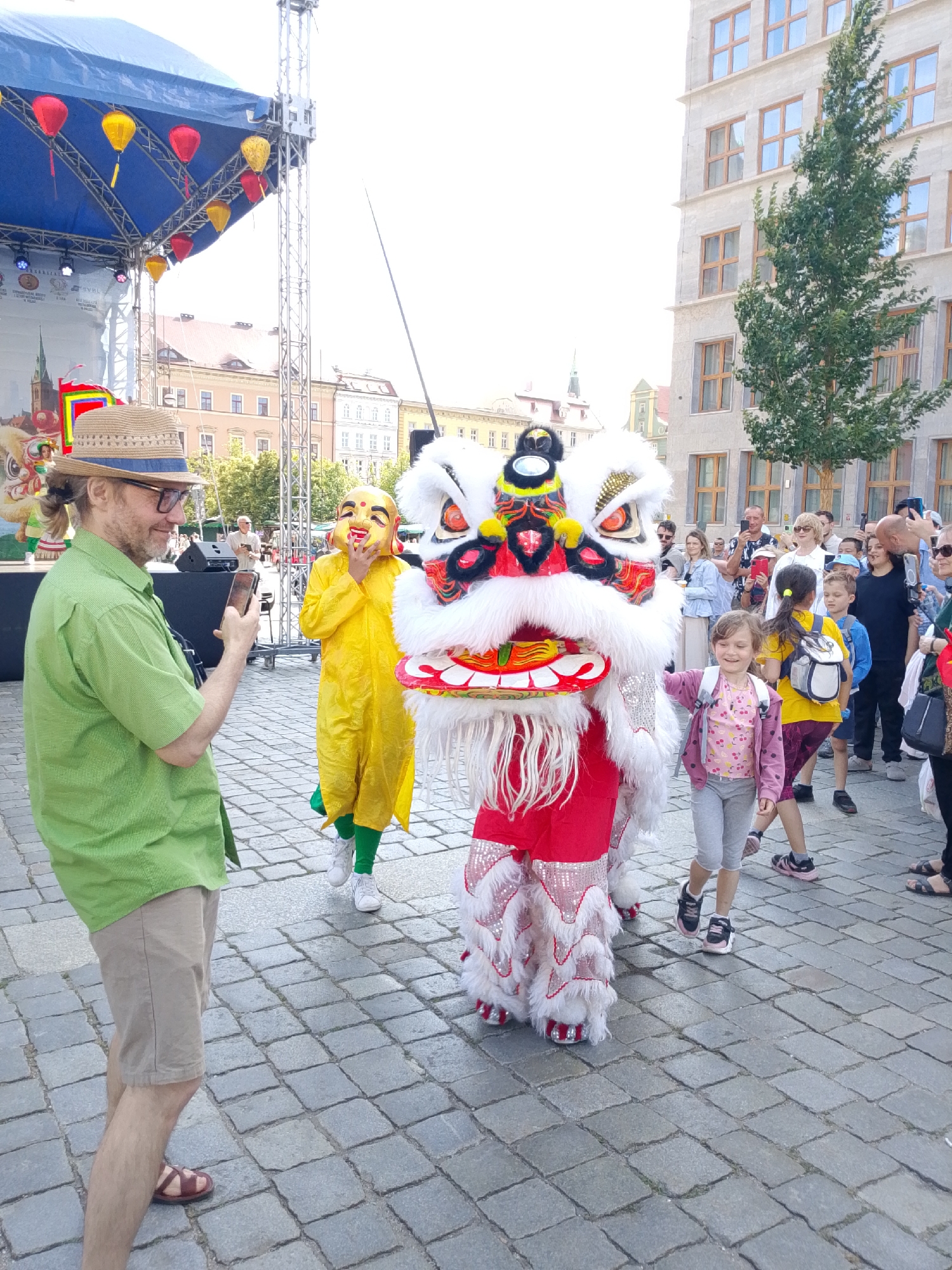 A street festival scene in Wrocław, Poland, features a person in a lion dance costume, composed of white fur and red embellishments, being paraded. Two individuals in yellow costumes with masks are near the lion, with one touching their mask. A man wearing a straw hat and glasses is photographing the event with his phone. In the background, a stage with a blue canopy is visible, decorated with red and yellow lanterns. A crowd of onlookers, including children and adults, are gathered to watch the performance. The event takes place on a cobblestone street in front of buildings with classical architecture.