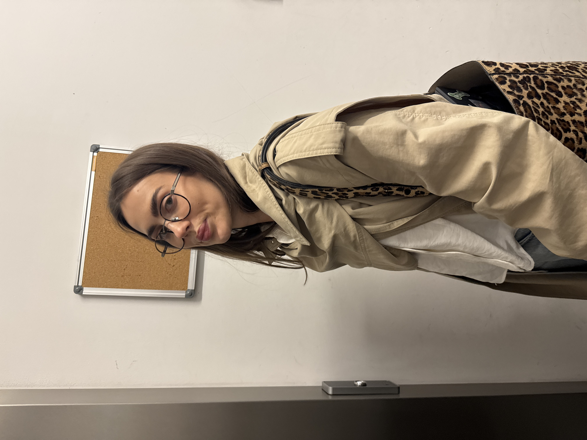 A woman with long brown hair and glasses leans into the frame, wearing a tan trench coat and a leopard-print bag. A corkboard hangs on a white wall behind her. A dark countertop is visible in the foreground, with a silver object resting on it. The location is Unknown, Unknown.