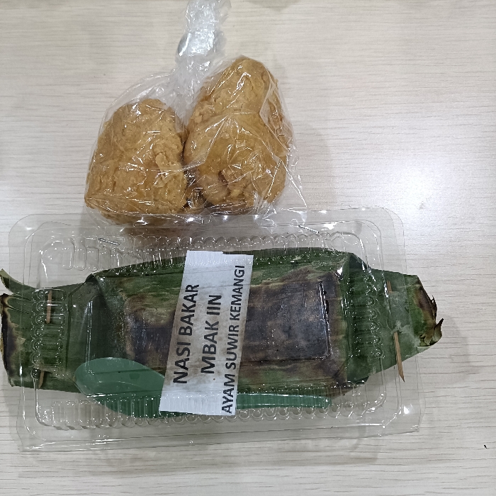 A plastic container with a green banana leaf inside holds a wrapped food item on a wooden surface. The container has a sticker on it which reads "Nasi Bakar Mbak Iin Ayam Suwir Kemangi". Two wrapped, fried, food items are to the left of the container on a white plastic bag. The image appears to be taken in a restaurant or food vendor in Tugu, Indonesia, with coordinates -6.973, 110.326. The image shows a close-up of the food items and the container, with the surrounding environment out of focus. The food appears to be Indonesian street food and is ready to be eaten.