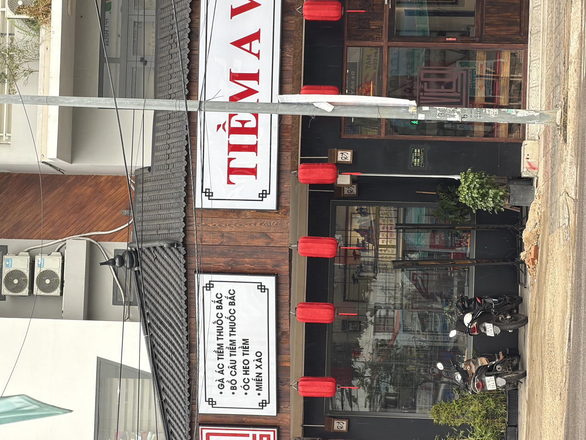 The image shows a street view in Nha Trang, Vietnam. The primary focus is on the facade of a building housing a shop. The shop has a dark-colored wooden exterior and features large glass windows, through which interior displays and furnishings are partially visible. Two red lanterns hang from the eaves of the shop, adding a decorative element. Above the windows, a white sign with red Vietnamese text, likely the shop's name or offerings, is displayed. Another smaller white sign with bullet points lists services such as "Gà ác tiêm thuốc bắc" (Black chicken herbal injection), "Bồ câu tiêm thuốc bắc" (Pigeon herbal injection), "Óc heo tiêm" (Pig brain injection), and "Miến xào" (Stir-fried noodles).
In front of the shop, two motorbikes are parked on the sidewalk. One is brown and black with a license plate reading "79-N2 164.07". The other is black with a license plate reading "79-CT 31R.25". Green potted plants are placed along the sidewalk, one large one to the left of the motorbikes and a smaller one to the right of the shop entrance. A street light pole stands prominently in the foreground, partially obscuring the shop's signage. The street itself appears to be paved, with some construction debris visible on the right side of the image. The building behind the shop is lighter in color, with visible air conditioning units mounted on its exterior. The sky is overcast, suggesting a cloudy day.