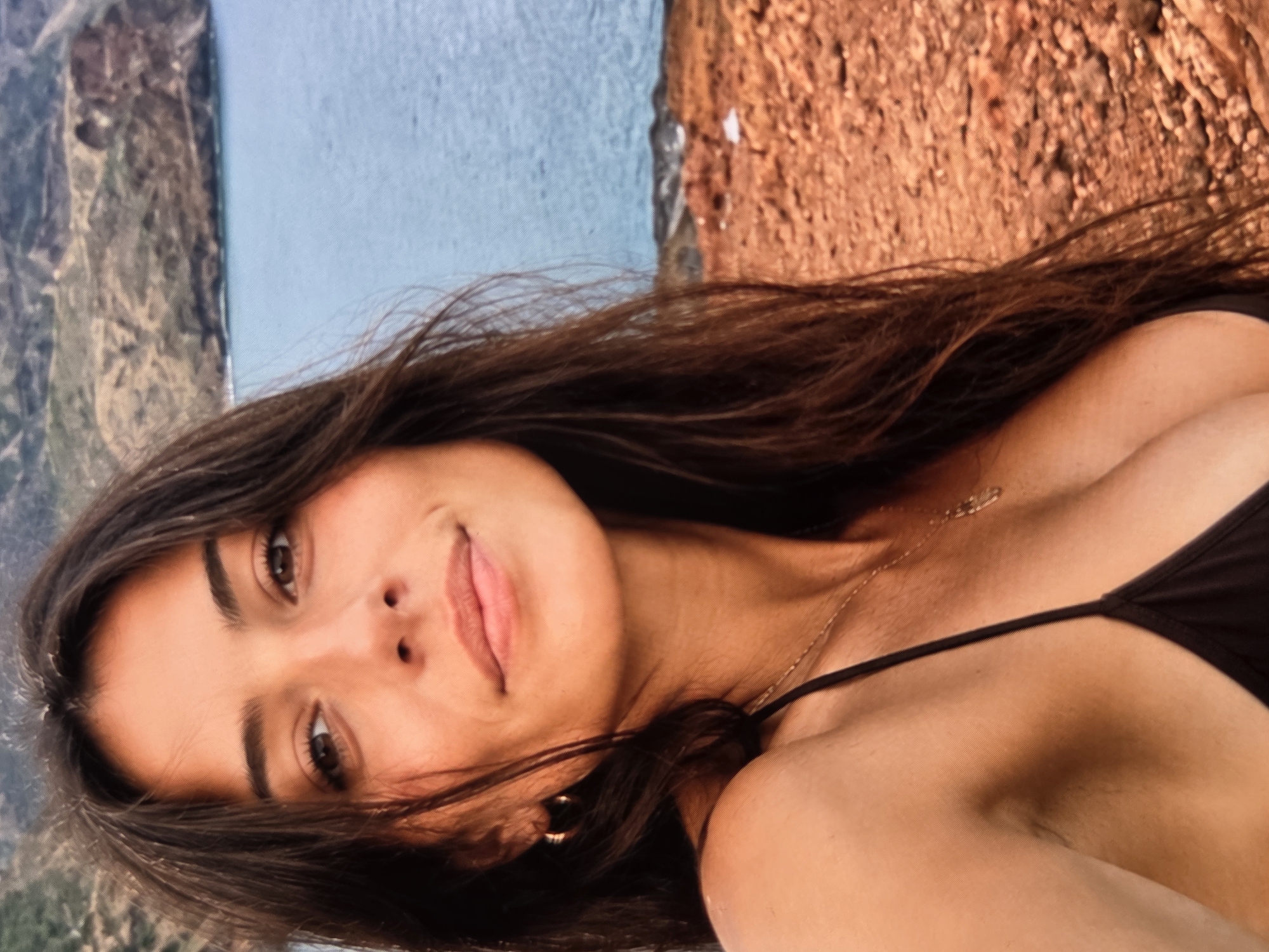 A medium shot features a woman with brown hair, brown eyes, and a light complexion, posing against an outdoor backdrop. She is wearing a thin-strapped black top and a gold necklace. The background consists of a body of water on the left and a rough, textured surface, possibly rock or earth, on the right. The photo appears to have been taken from a high vantage point.
The location cannot be determined to be Sofia, Bulgaria from the image provided.