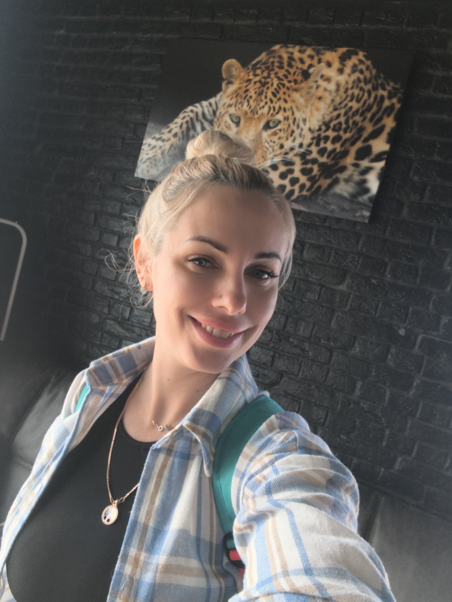 A woman is taking a selfie indoors. She has blonde hair in a bun, fair skin, and is smiling. She is wearing a black top, a blue and white plaid shirt, and a gold necklace. Behind her is a black brick wall with a picture of a leopard hanging on it. The location may be Орджоникидзевский поселковый совет, Ukraine, but there is no explicit indication of this in the image. The woman is interacting with her phone to take the selfie.