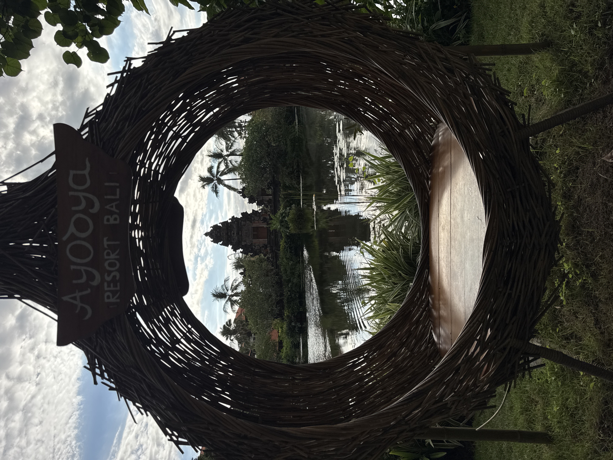 The image captures a view through a circular structure made of woven twigs at the Ayodya Resort Bali in Kuta Selatan, Indonesia. A wooden bench is integrated into the structure. The view through the opening reveals a tranquil lake surrounded by tropical vegetation, including palm trees. A Balinese temple is visible on the far side of the lake. The lake's surface reflects the temple, trees, and sky, which is partly cloudy. A sign bearing the words "Ayodya Resort Bali" is on the side of the woven twig structure. The surrounding area is grassy, and there are plants on either side of the structure.