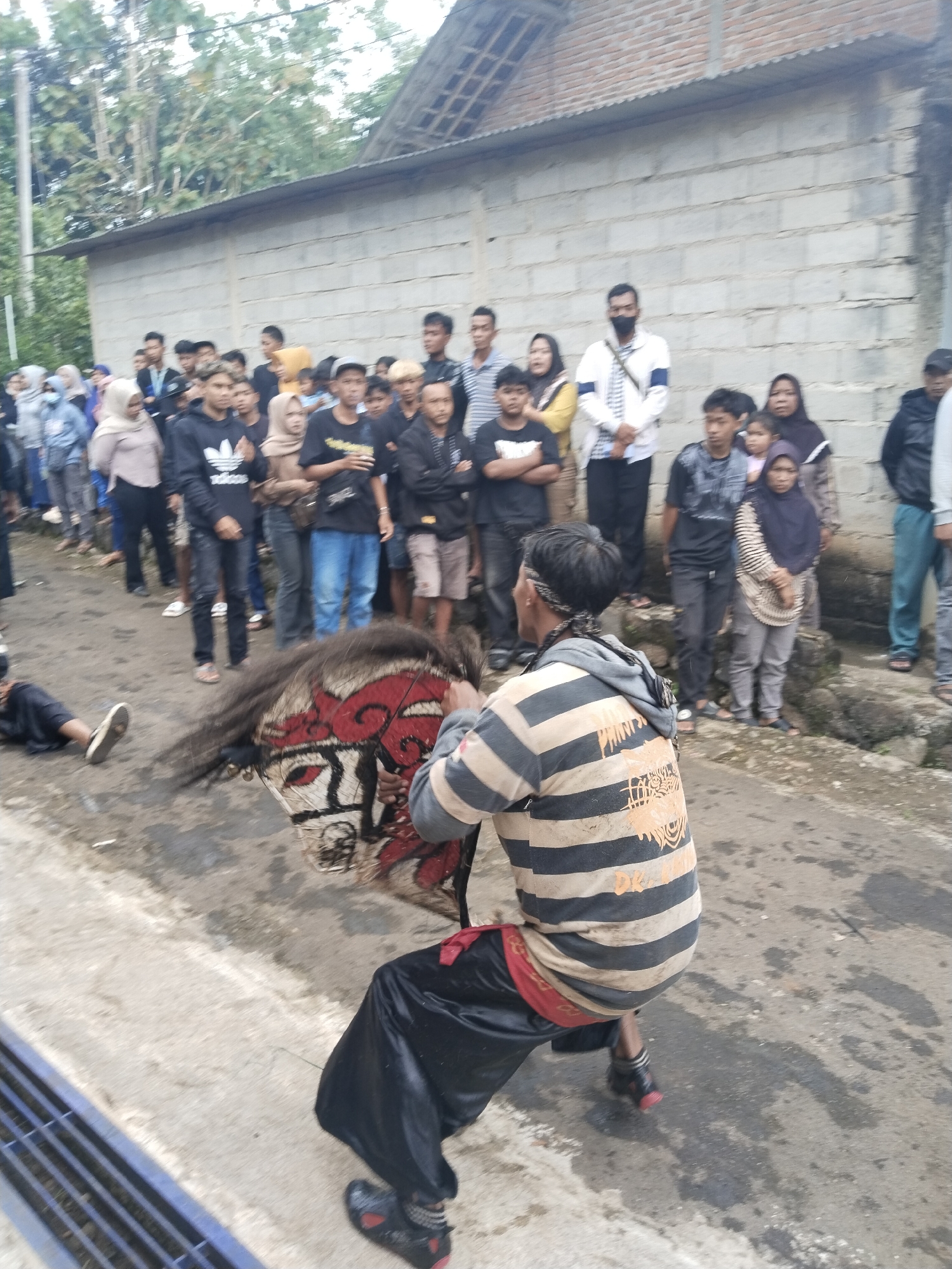 In Pendem, Indonesia, a crowd of people observes a man in a horizontally striped tan and black shirt, black pants, and red accents, squatting with a traditional horse effigy. The effigy is predominantly red, white, and black. A person lies prone on the ground to the left of the performer. A concrete block wall runs along the back of the crowd. The sky is overcast, and the street appears wet.
