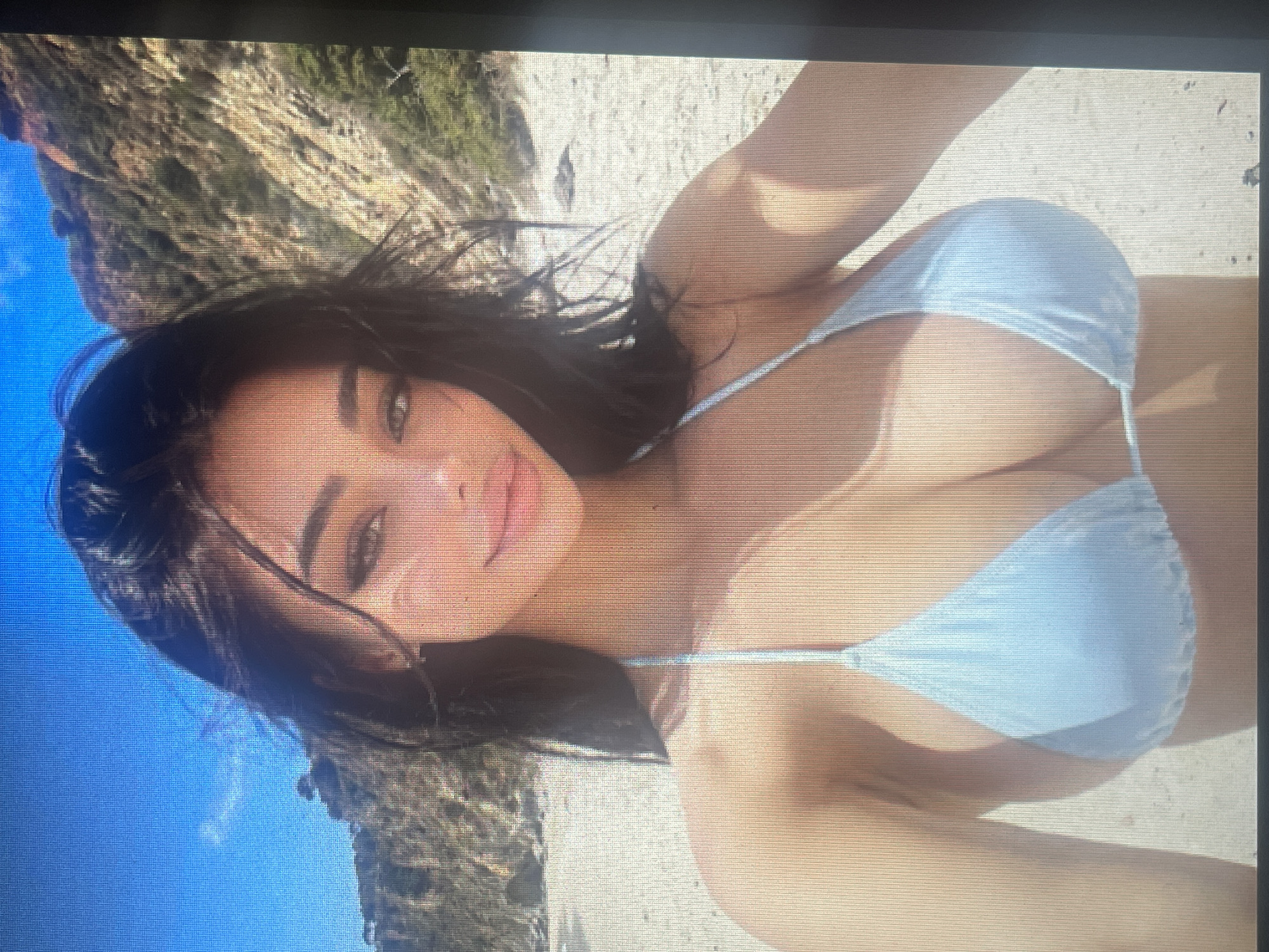 A young woman with dark hair and light skin is posing for a selfie on a sandy beach. She is wearing a light blue bikini top. Behind her, there is a hillside with sparse vegetation and a clear blue sky. The image is taken from the chest up. There is no indication of the city of Mantov, Czechia in this image.