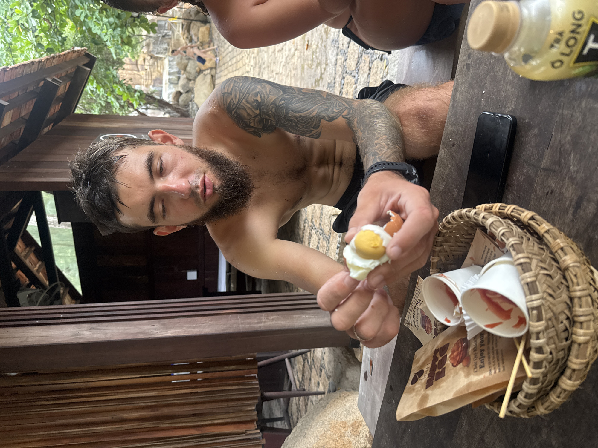 A man with a beard and tattoos is sitting at a wooden table, peeling a hard-boiled egg. He is shirtless, wearing black shorts. In front of him is a woven basket containing a brown paper bag, two white paper cups with red contents, and two wooden skewers. A smartphone and a bottle of O Long tea are also on the table. In the background, a person in a bikini is visible, and trees and rocks can be seen. The scene appears to be an outdoor shaded area, possibly near a beach or natural attraction. The presence of a souvenir bag suggests this may be a tourist location. The man's wet skin and the light clothing indicate a warm climate, consistent with Nha Trang, Vietnam.