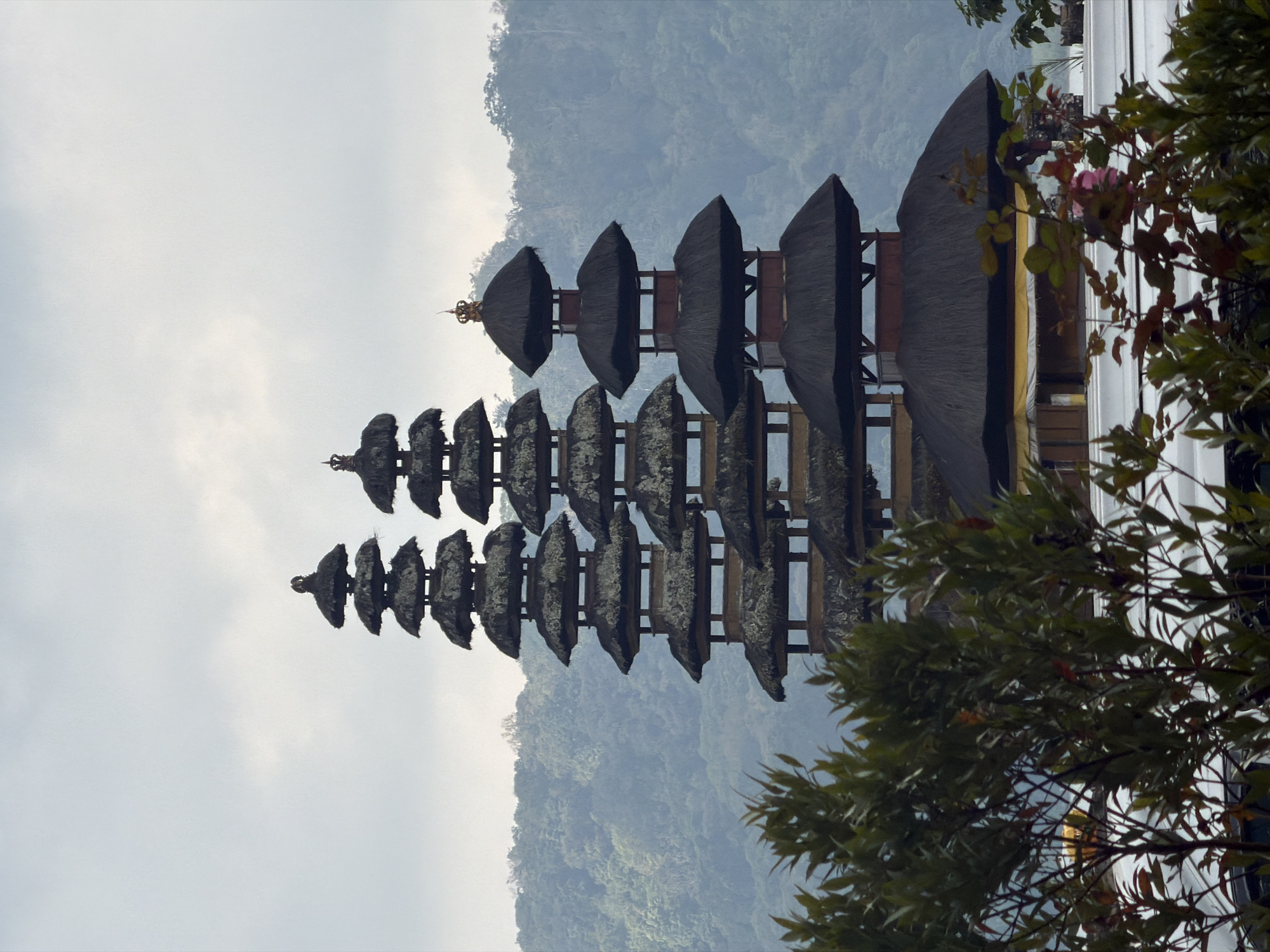 This image shows the Pura Ulun Danu Beratan temple complex, located on the shores of Lake Beratan in Bali, Indonesia, near the city of Banjar. The temple is characterized by its multi-tiered meru towers, which have thatched roofs and are designed to represent the different levels of the cosmos. The foreground is partially obscured by lush green foliage and some flowering plants, with a hint of white structures and possibly vehicles beneath them. In the background, rolling hills covered in dense, verdant forest recede into a hazy, overcast sky. The overall atmosphere is serene and somewhat muted due to the cloudy weather. No people are visible in the frame.
