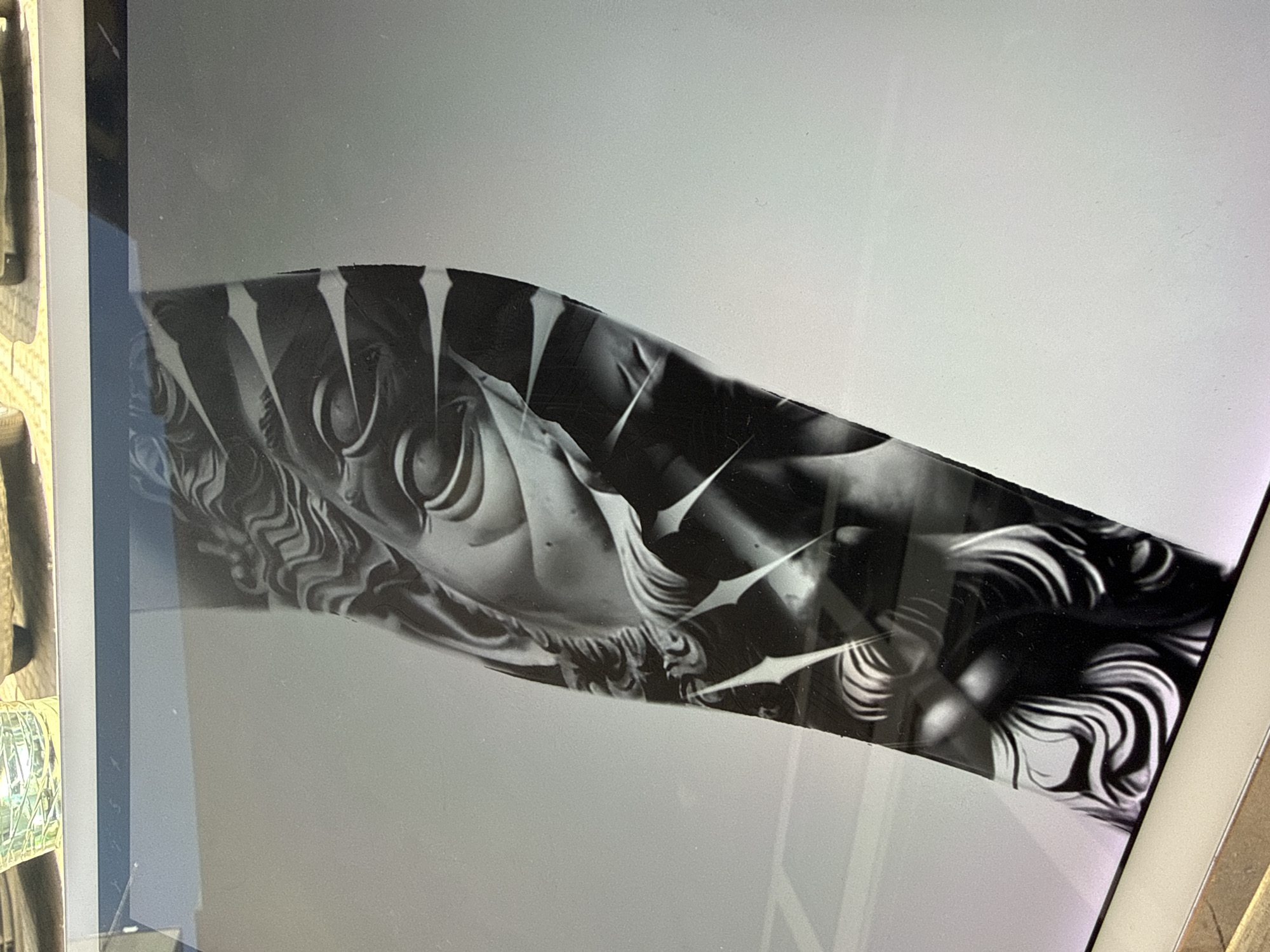 The image is a photograph of a tablet screen displaying a black and white image of what appears to be a tattoo design. The design includes detailed line work and shading, featuring a face with prominent eyes and stylized hair or feathers. Sharp, white geometric shapes are interspersed throughout the design. The tablet is situated within a cluttered environment, as a portion of a tray filled with small, unidentified objects is visible on the left edge of the frame. The location is not discernible from the image, and there is no indication it is in Sofia, Bulgaria.