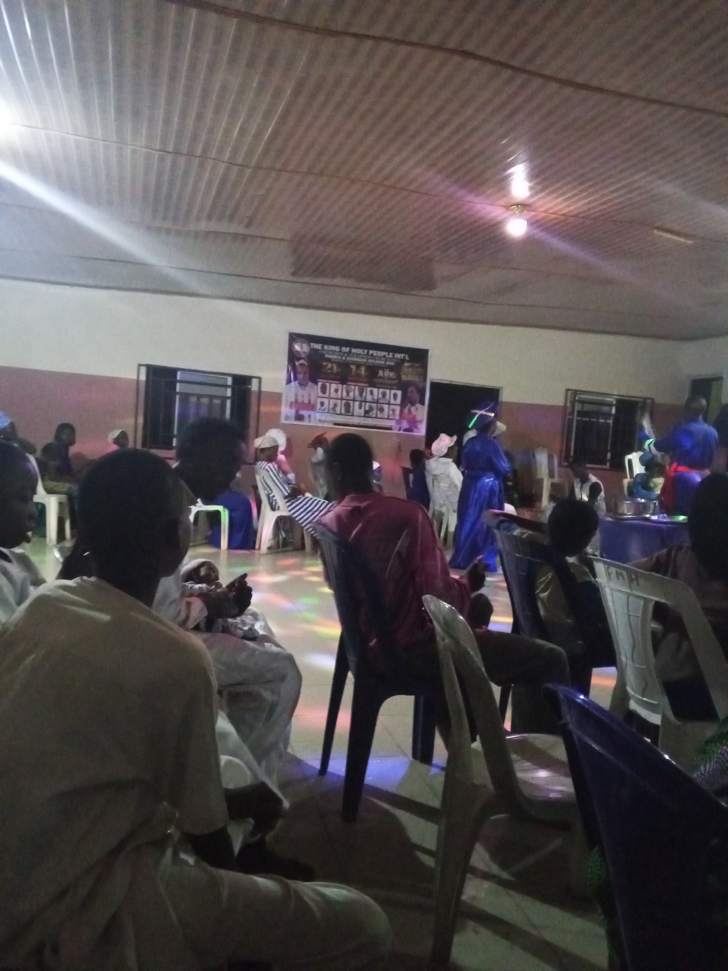 The image shows an indoor gathering in Lokoja, Nigeria. People are seated on plastic chairs, facing a stage area. The room has a corrugated metal ceiling with fluorescent lighting. A banner reading "The King of Holy People Int'l" hangs on the wall. Some individuals are wearing blue gowns. Colored lights are visible, creating a festive atmosphere.