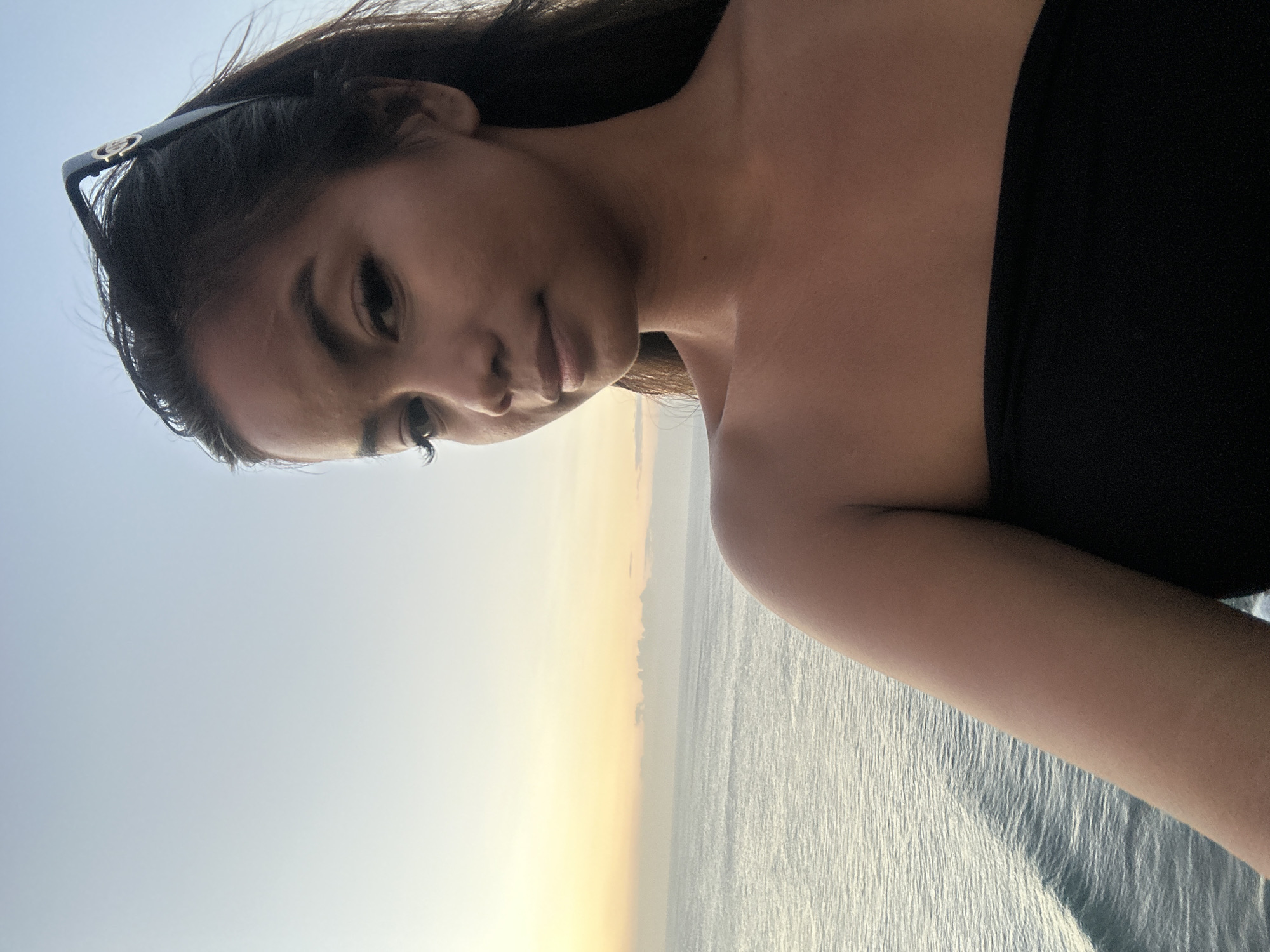 A young woman with long dark hair and sunglasses perched on her head is captured in a selfie, with a tranquil ocean and a pale orange sunset in the background. She is wearing a black strapless top. The scene appears to be Kuta Selatan, Indonesia, a popular tourist area known for its beautiful beaches and surf spots.