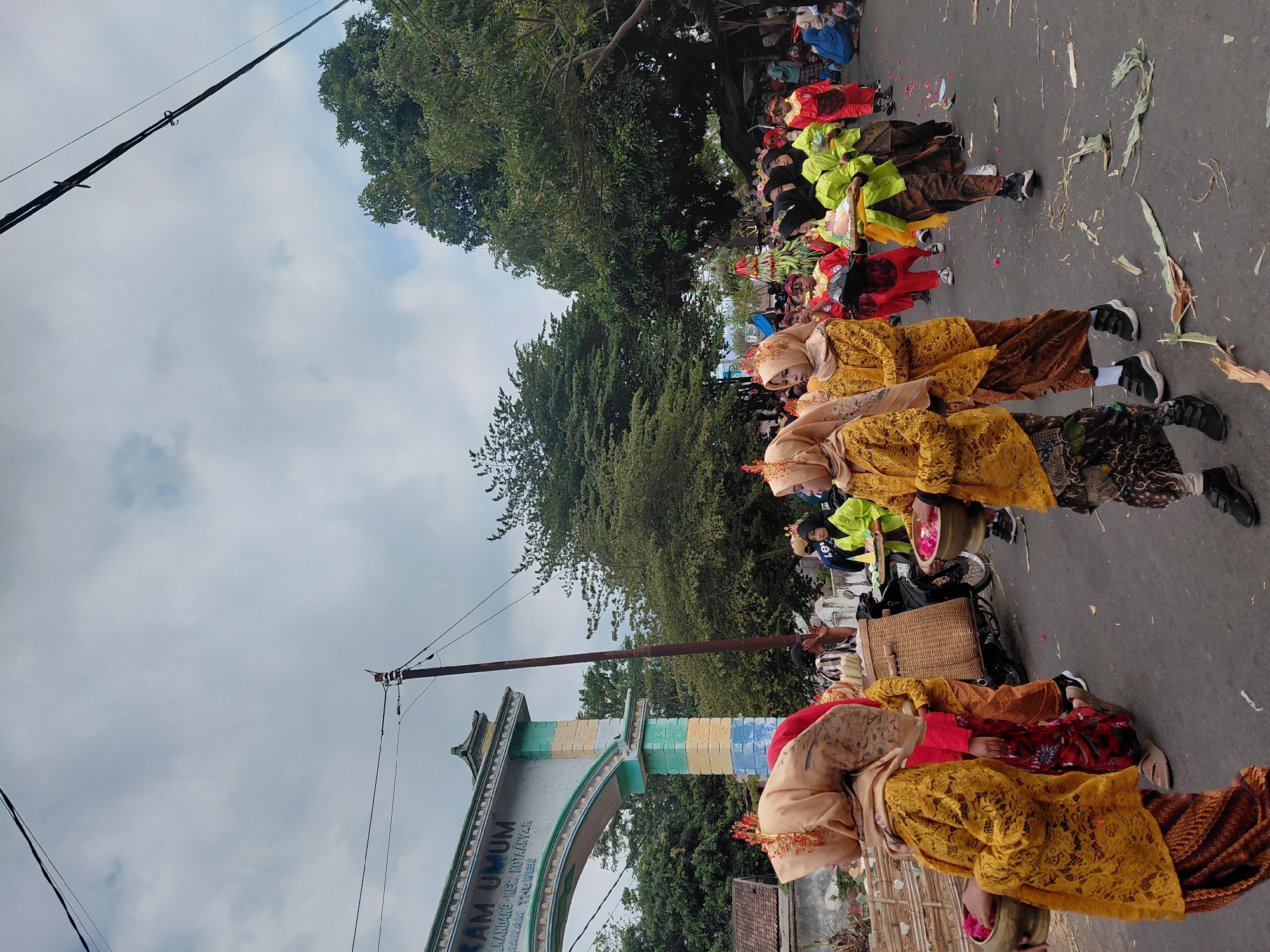 A group of children in traditional Indonesian attire, likely for a festival or cultural event, are marching down a paved street in Bimo, Indonesia. The children are wearing brightly colored garments, primarily yellow kebaya tops and patterned sarongs, with headscarves. Some are holding bowls filled with flowers, while others are carrying elaborate offerings or decorations.
In the background, an archway with inscriptions in Indonesian suggests a formal entrance to a neighborhood or institution. Trees line the street, and a cloudy sky looms overhead. Several adults are present, some observing the procession and others participating. A motorcycle with a large wicker basket is parked nearby. The street is adorned with scattered flower petals and fallen leaves, indicating the ongoing festivities. The overall scene captures a vibrant community celebration within an Indonesian setting.