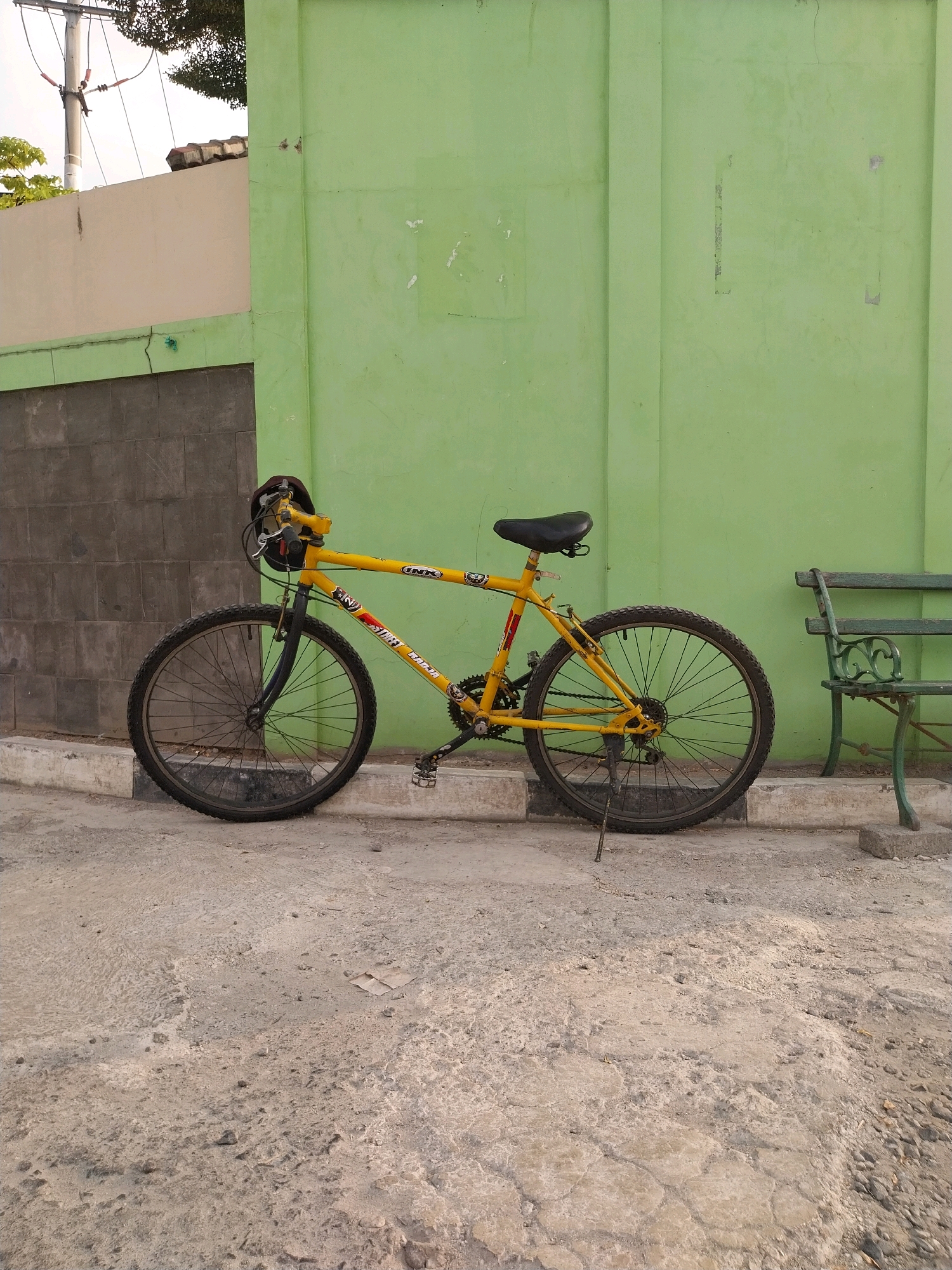 A bright yellow mountain bike with black tires and a black seat is parked on a textured concrete surface next to a light green wall. The bike has a red and white decal on its frame and a black helmet resting on the handlebars. To the right of the bike, a dark green metal bench with ornate detailing is also present. Behind the bike and bench is a wall, partially covered by a dark grey tiled section. A lighter colored wall with a tiled roof is visible in the background on the left, with trees and power lines suggesting an outdoor urban setting. This scene is located in the city of Cirebon, Indonesia.