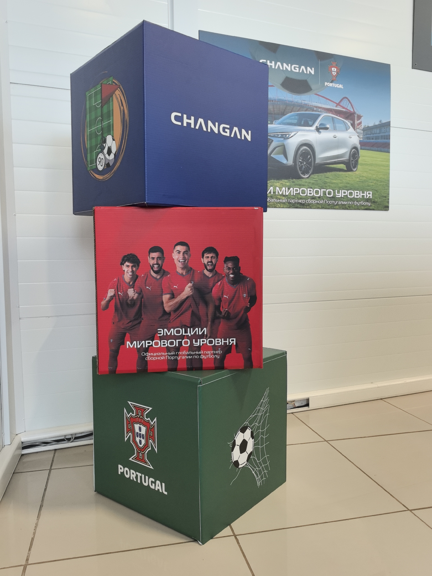 The image shows a display of three stacked cardboard boxes, presumably for a promotional event. The top box is dark blue with "CHANGAN" written on one side and a graphic of a soccer field, a soccer ball with a stopwatch showing "90," and a flag on another side. A poster is partially visible behind this box, featuring a silver car, the "CHANGAN" logo, and "PORTUGAL" with the Portuguese national football team crest. The middle box is red and displays a photograph of four soccer players in red jerseys, with the text "EMOTIONS OF THE WORLD LEVEL" and "Official global partner of the Portugal national football team" in Russian. The bottom box is green and features the Portuguese national football team crest and "PORTUGAL" on one side, and a soccer ball in a net on another side. The boxes are arranged in a staggered manner. The background consists of a white, ribbed wall and a tiled floor. The lighting suggests an indoor setting, possibly a showroom or event space. There is no indication of the location being in Perm, Russia.