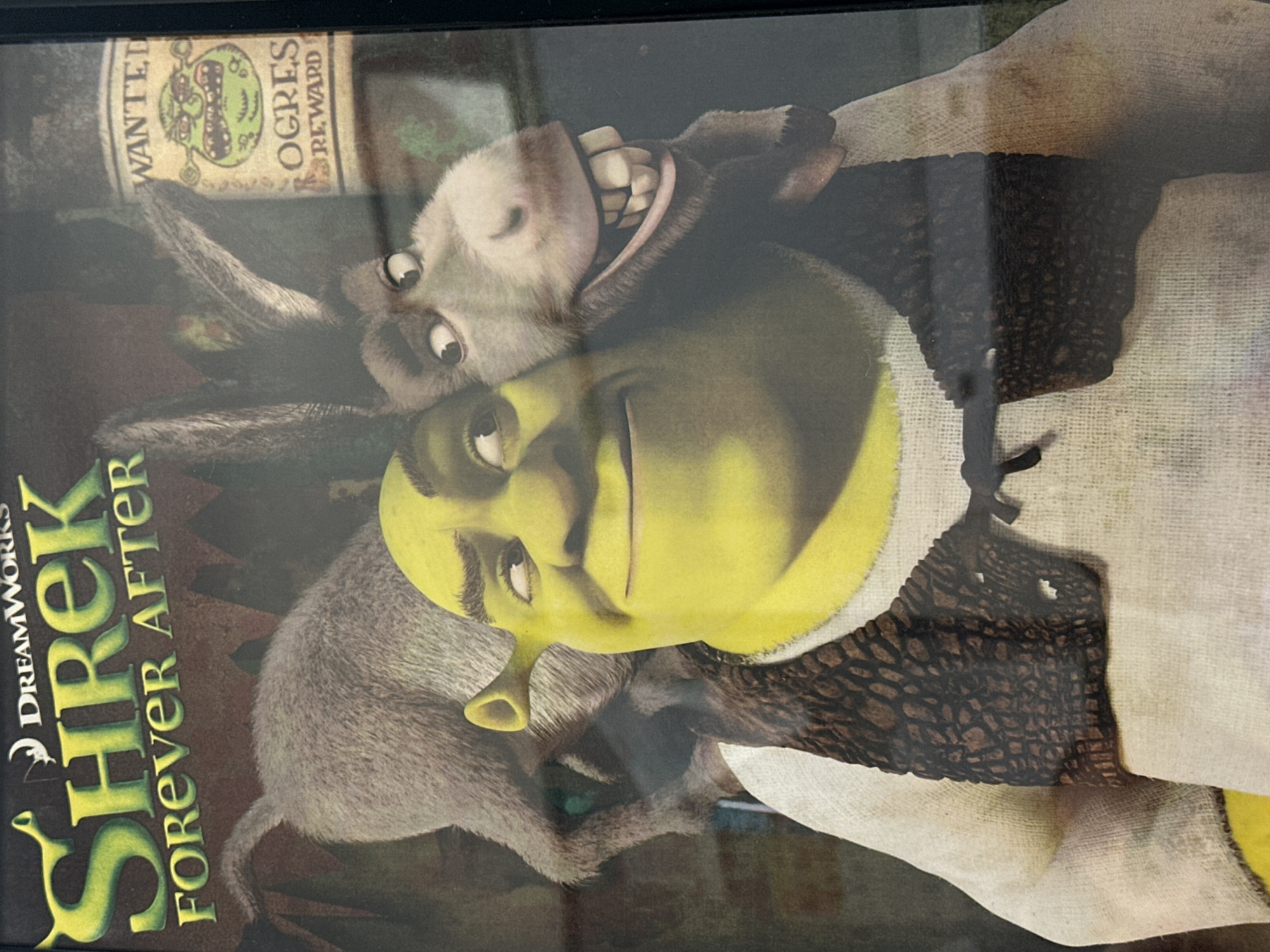 The image shows the DVD cover for the movie "Shrek Forever After." In the foreground, Shrek, an ogre with green skin and a large head, is wearing a white tunic and a brown vest. His right ear is visible, sticking out from the side of his head. Behind Shrek, Donkey, a gray donkey with large ears, is nuzzling Shrek's head. Donkey's mouth is open, revealing his teeth. In the background, a "Wanted" poster for "Ogres" is visible on the right side of the image. The movie title "Shrek Forever After" is displayed at the top of the cover. This image is a close-up of the DVD cover and does not depict a scene in the city of Harrisburg, United States of America.