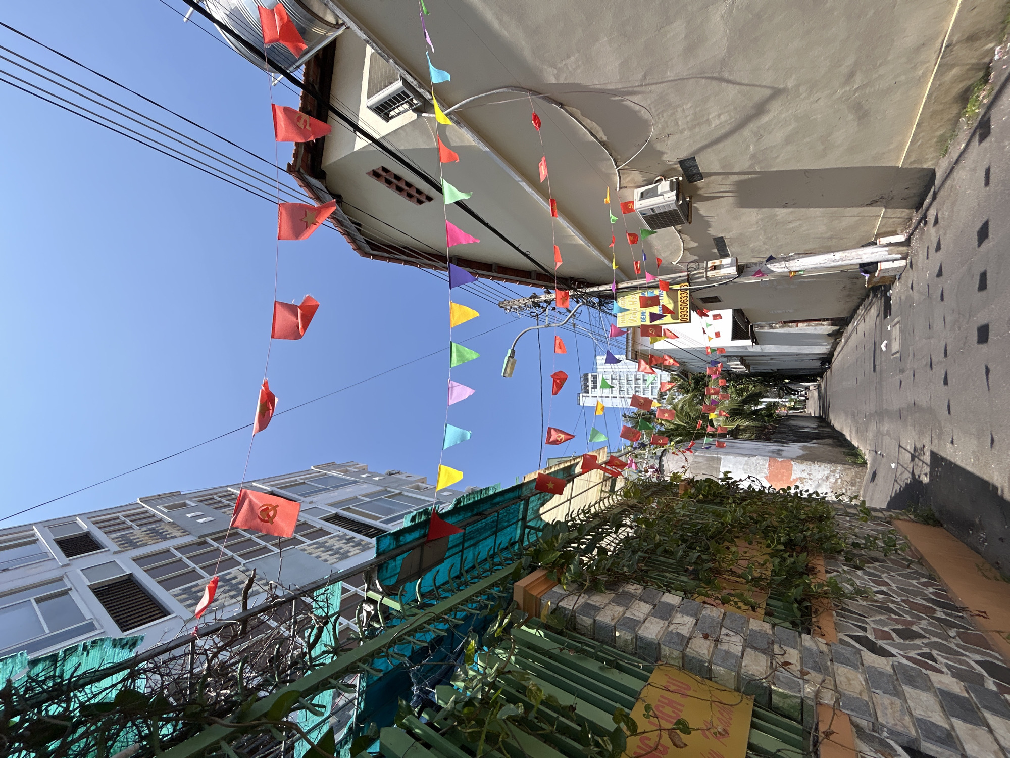The image shows a narrow alleyway in what appears to be Bắc Nha Trang, Vietnam, judging by the flags displayed. The sky is clear and blue. Buildings line both sides of the alley. Banners with colorful triangular flags and small red flags with a yellow star are strung across the alleyway, overhead. Power lines also run along the top of the alley. On the left, there is a building with many windows and green coverings. Below, there is a green fence with some vines. On the right is a wall constructed of stone. The alley appears to lead toward a building in the distance. There are no visible people in the immediate scene.