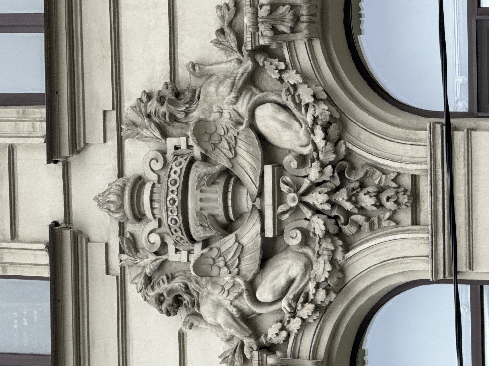 Architectural detail on a building in Saint Petersburg, Russia. Features carved stone elements including lions with wings flanking a central urn-like motif. Foliage and decorative scrollwork surround the composition. A round window is visible to the right. The building facade is a light color, suggesting stone or stucco.