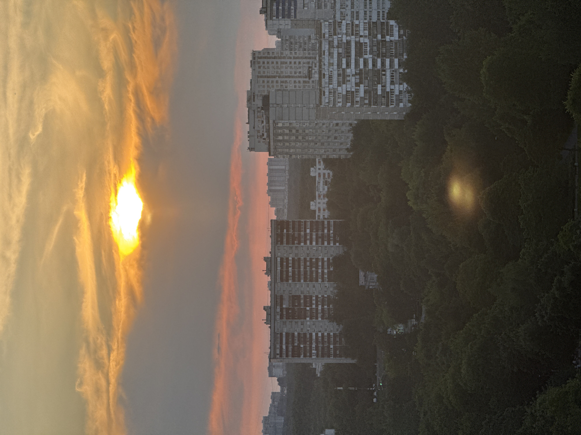 The image depicts a panoramic view of a residential area in Moscow, Russia, during sunset. The sky is filled with dramatic clouds, ranging from bright yellow and orange near the setting sun to softer pinks and grays further away. The sun itself is a brilliant, almost white orb, partially obscured by clouds.
Below the sky, a dense canopy of dark green trees dominates the middle ground, interspersed with the tops of various residential buildings. These buildings are typical Soviet-era panel apartment blocks, characterized by their uniform facades, numerous balconies, and modular construction. They vary in height and design, with some featuring lighter-colored panels and others darker, weathered concrete. One prominent building in the center is a multi-story block with a grid-like pattern of windows and balconies.
In the background, more apartment buildings are visible, some appearing further away and shrouded in the hazy twilight. A few streetlights or illuminated windows can be seen within the residential blocks and amidst the trees, suggesting urban activity. The overall impression is one of a densely populated urban landscape at the end of the day, with nature and architecture coexisting. There are no identifiable people or specific actions depicted in the scene. The atmosphere is tranquil, with the sunset casting a warm glow over the city.