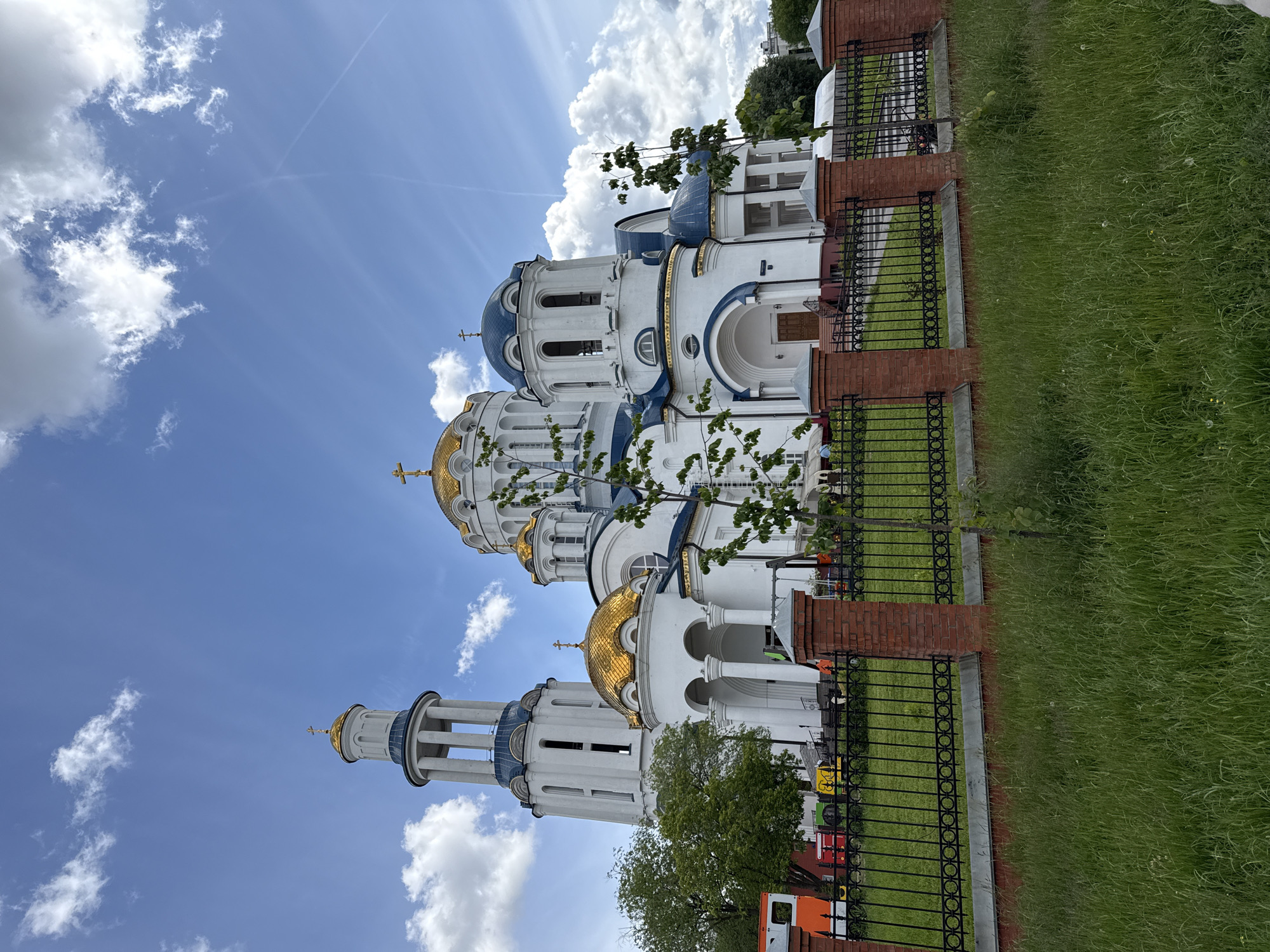 The image shows a white cathedral with golden and blue domes, likely an Orthodox church, located in Moscow, Russia. The building is surrounded by a wrought iron fence atop a brick base. There is a green lawn in front of the fence. The sky is blue with scattered white clouds. Several vehicles (orange, yellow, red, green) are visible behind the fence and a small tree is in the foreground. The photograph is taken from a low angle, emphasizing the height of the church.