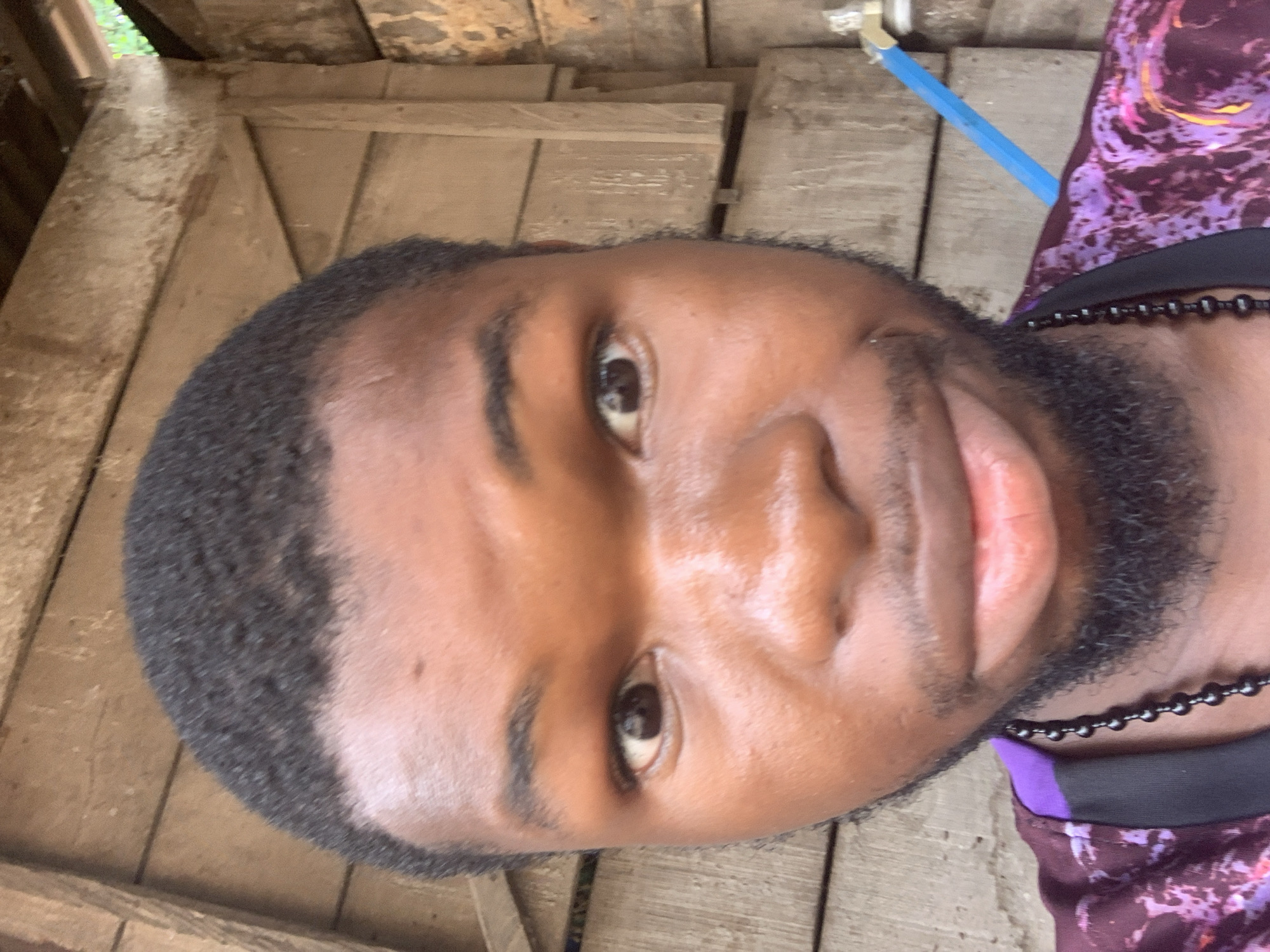 A close-up, slightly angled shot focuses on the face of a dark-skinned man with a short, dark haircut and a neatly trimmed beard. He is wearing a dark beaded necklace and a purple and white patterned shirt. The background consists of weathered wooden boards, possibly a wall or floor, and a blue pipe is also visible. It's difficult to determine the specific location in Onitsha, Nigeria, from this image alone.