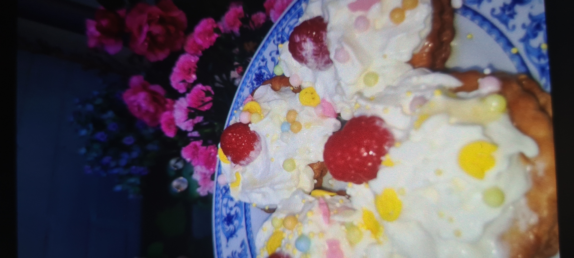 The image shows a close-up of several pastries topped with whipped cream, raspberries, and colorful sprinkles. The pastries are served on a plate with a blue and white patterned rim. In the background, a bouquet of vibrant pink flowers is visible. The lighting suggests it might be daytime, but the specific time and weather are not discernible. The setting appears to be indoors, possibly a home or a restaurant. There is no visible text in the image. The overall impression is one of a celebratory or dessert-focused occasion. The focus is on the details of the dessert, highlighting its festive appearance.