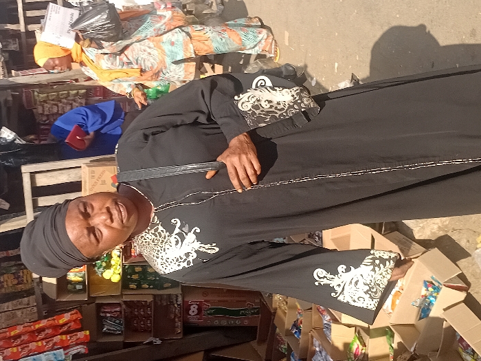 A woman in a black, decorative abaya and a black headwrap stands near an outdoor shop. Boxes of food items are visible. Behind her, another woman wearing a colorful patterned dress and a yellow head covering is resting on a makeshift bed. The setting appears to be a busy marketplace. The woman in the abaya is facing the camera.